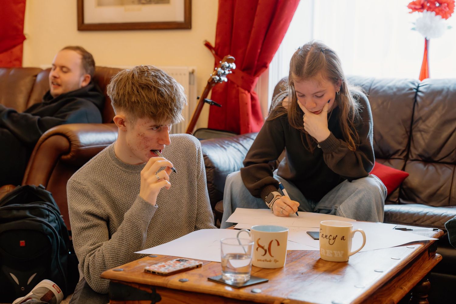 Two teenagers study at a wooden coffee table in a cozy living room with a guitar and red curtains in background.