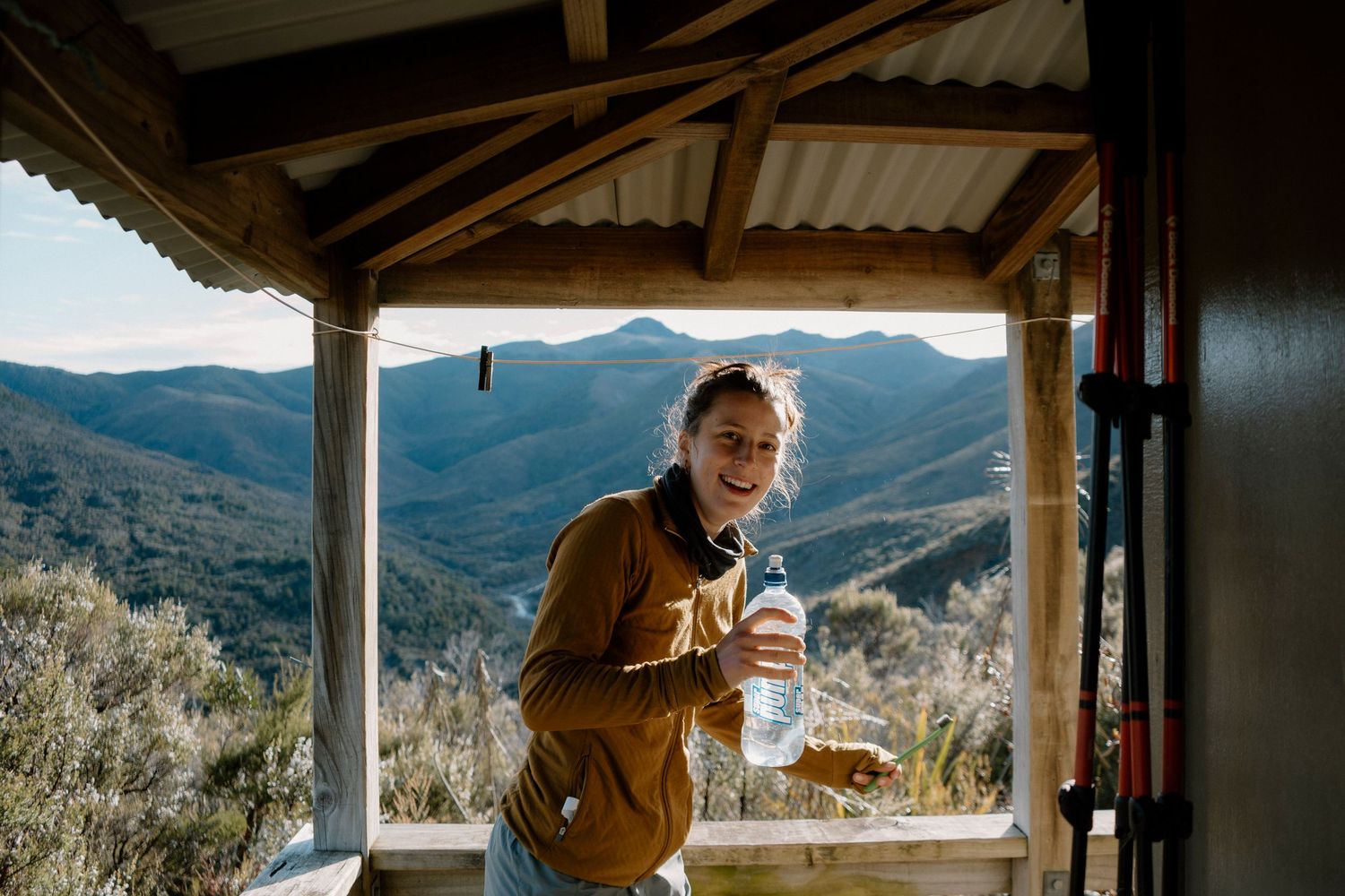 Person in brown jacket enjoys a hot beverage while looking at scenic mountain views from rustic cabin porch.