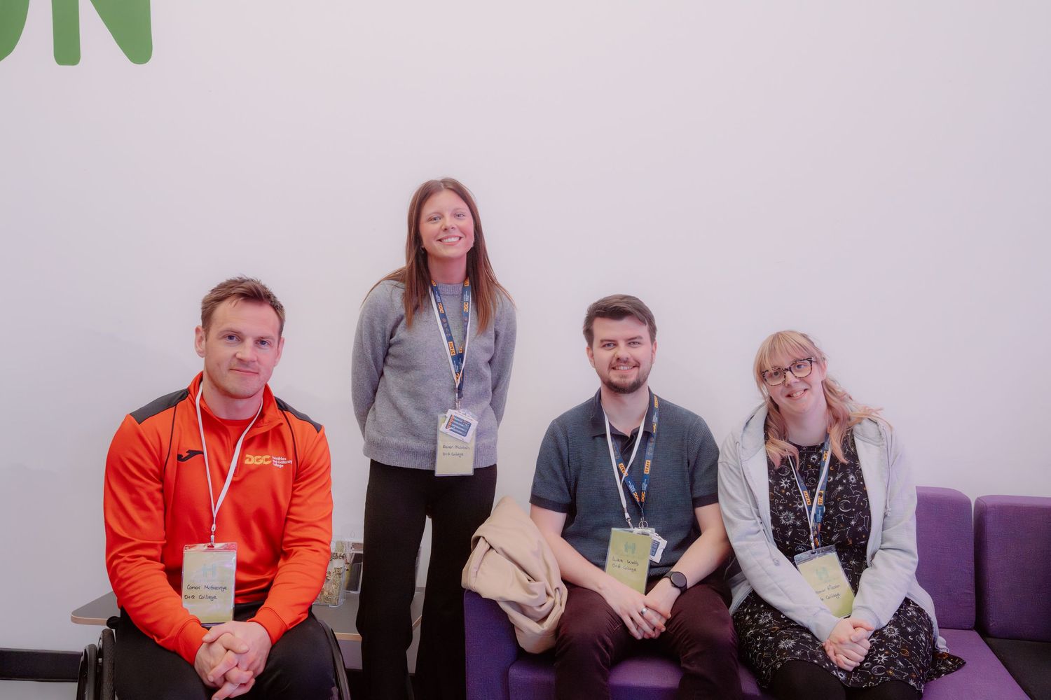 Four professionals wearing lanyards pose together smiling in a modern office space with purple seating.