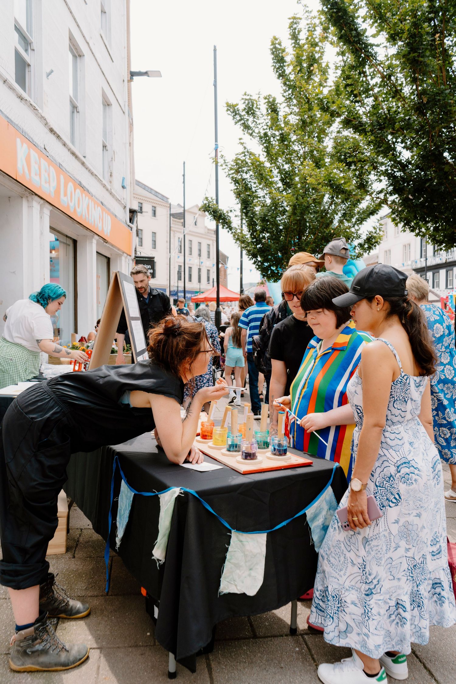 People browsing a street market stall with colorful drinks on a black table, outdoors on a busy urban sidewalk.