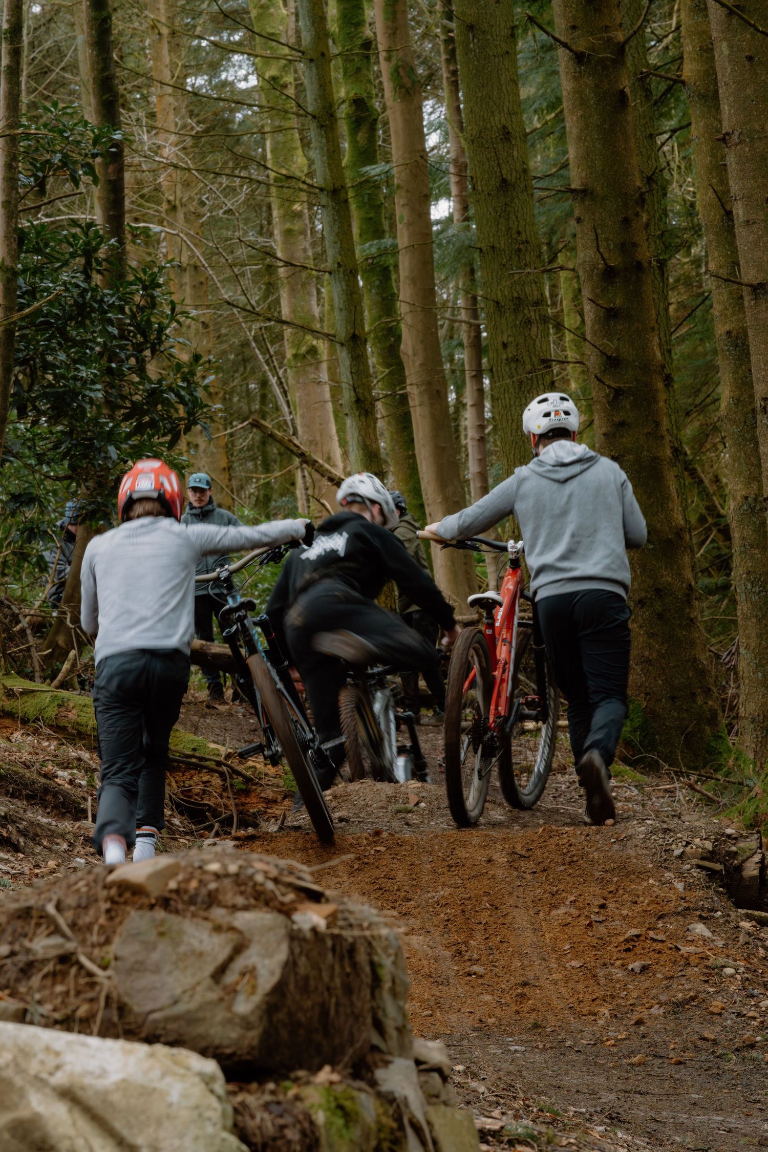 Group of mountain bikers with helmets pushing bikes up a dirt trail through a dense forest.