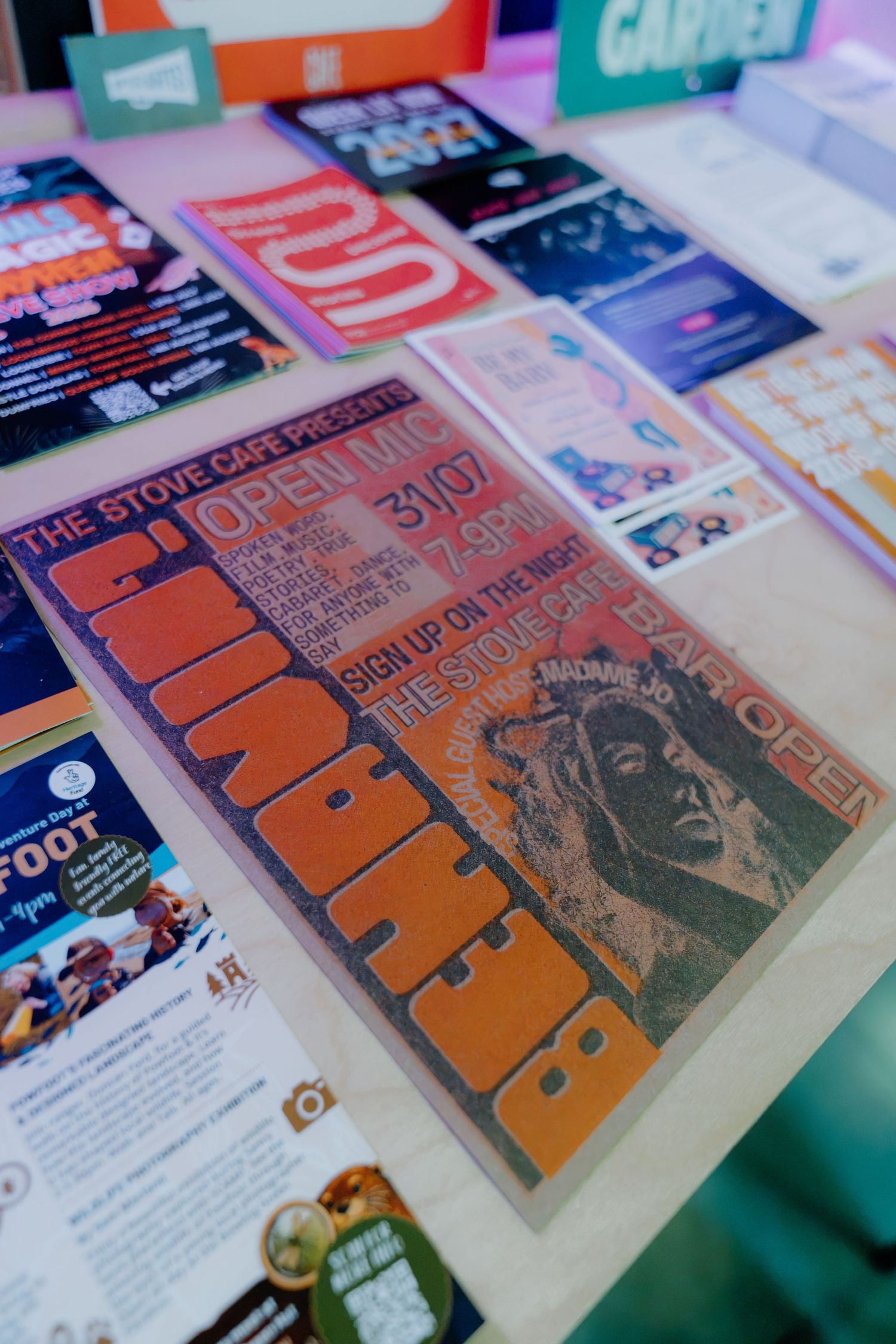 Colorful event flyers and posters spread on a table, featuring The Stove Cafe Open Mic night advertisement.
