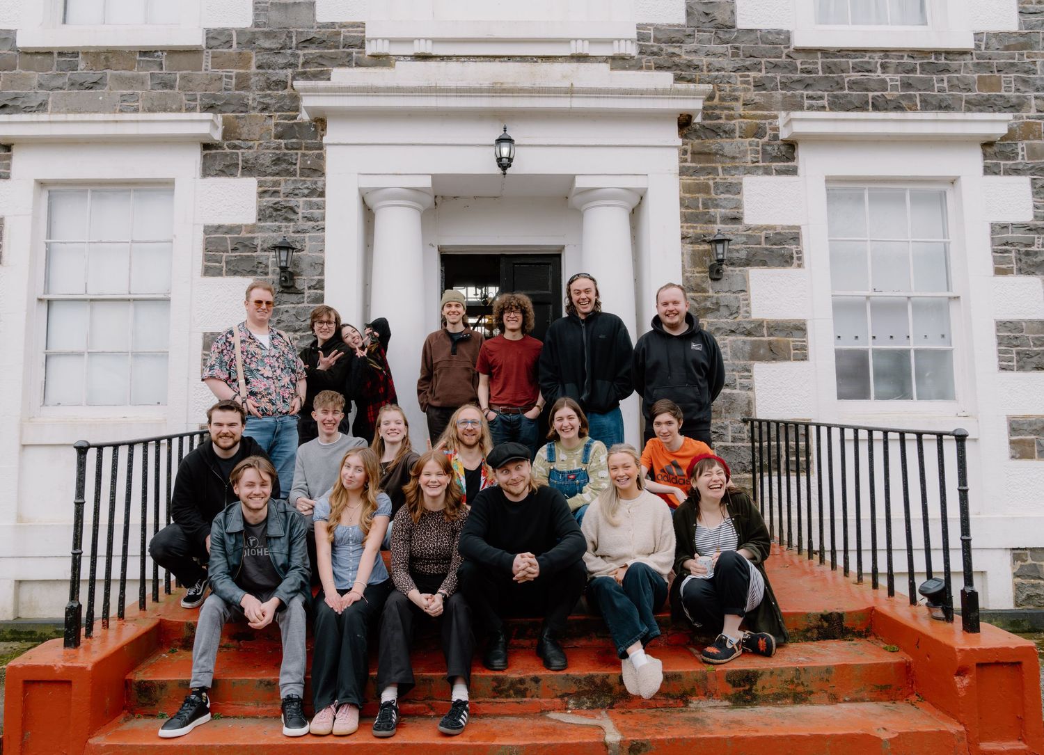 Group of young people posing on steps outside a historic stone building with white facade.