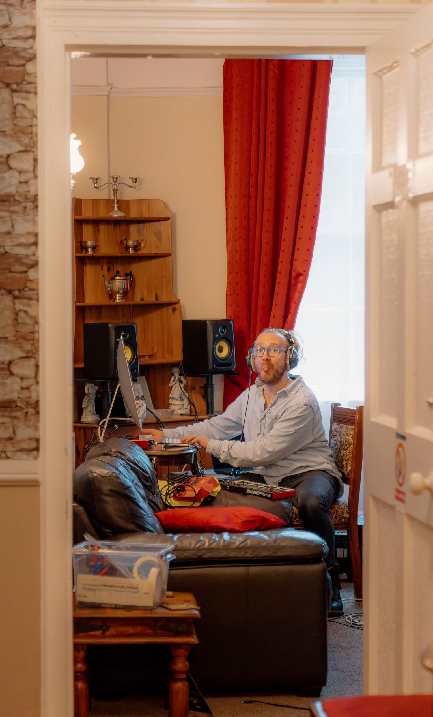 Man wearing headphones works at a home recording studio setup with monitor speakers and red curtains.