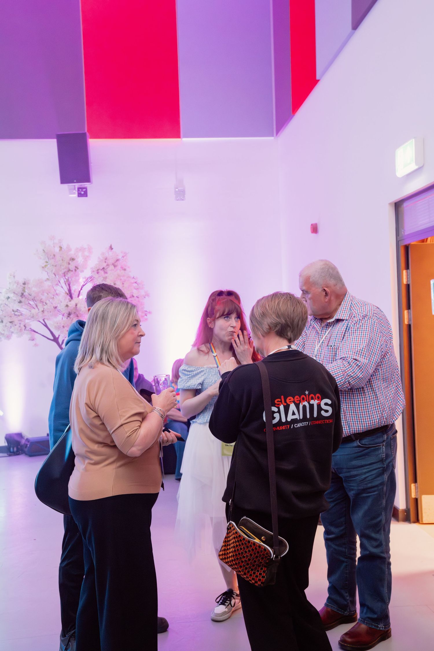 Group of people networking at an indoor event venue with pink lighting and decorative cherry blossom trees.