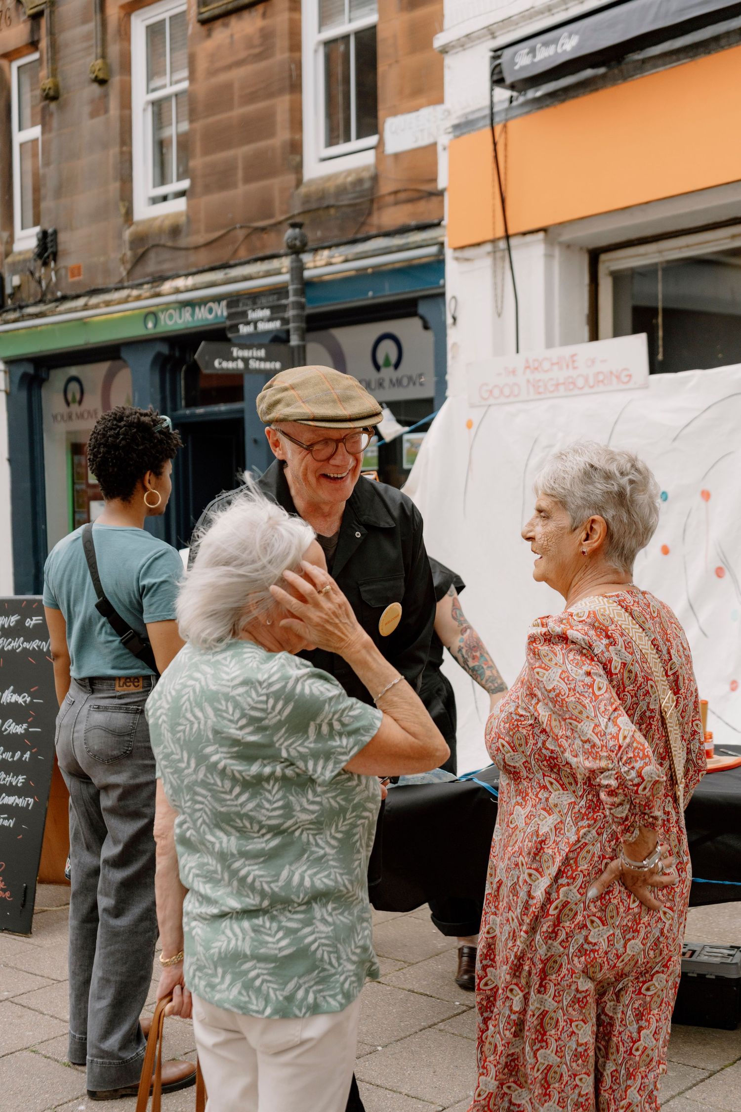 Elderly people laughing and chatting outdoors on a UK high street, with shops and a white van in the background.