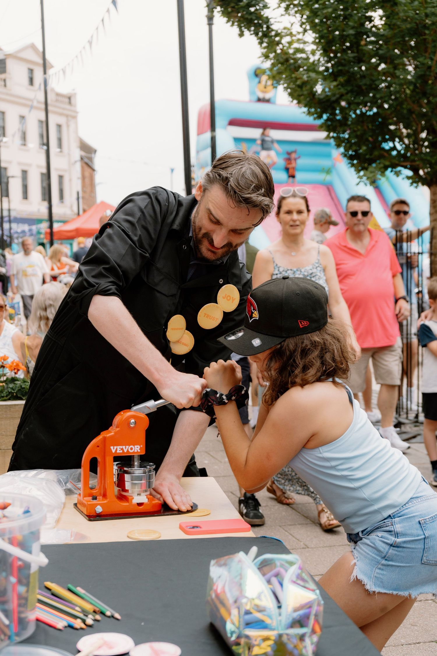 Man and girl use orange VEVOR badge press machine at outdoor festival table with colored pencils and bouncy castle in background.