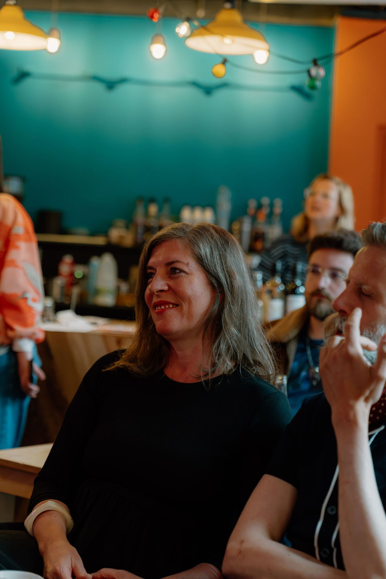 A smiling woman with gray hair in a black top enjoys a lively conversation at a colorful bar or cafe.