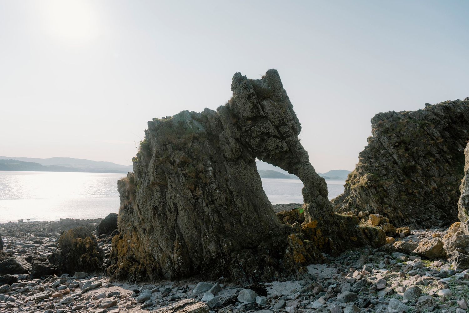 Natural rock arch formation along a rocky coastline with sunlit water and distant hills in the background.