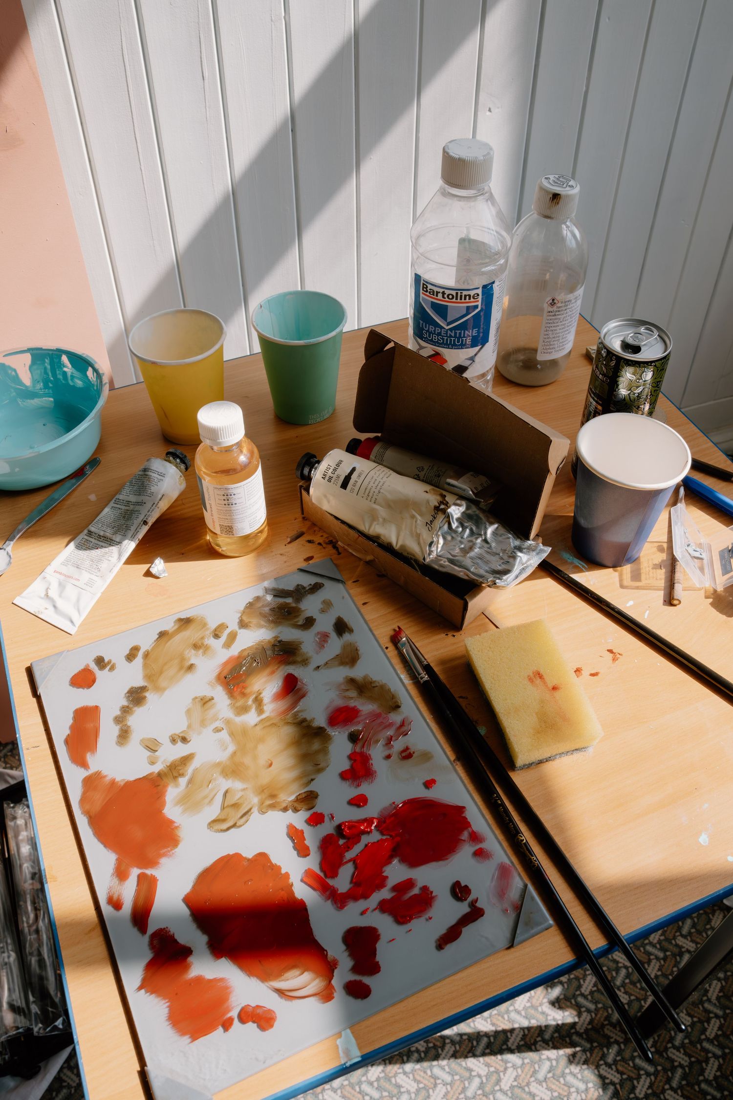 Artist's messy worktable with paint-stained palette, brushes, solvents, and colorful cups in a sunlit studio.