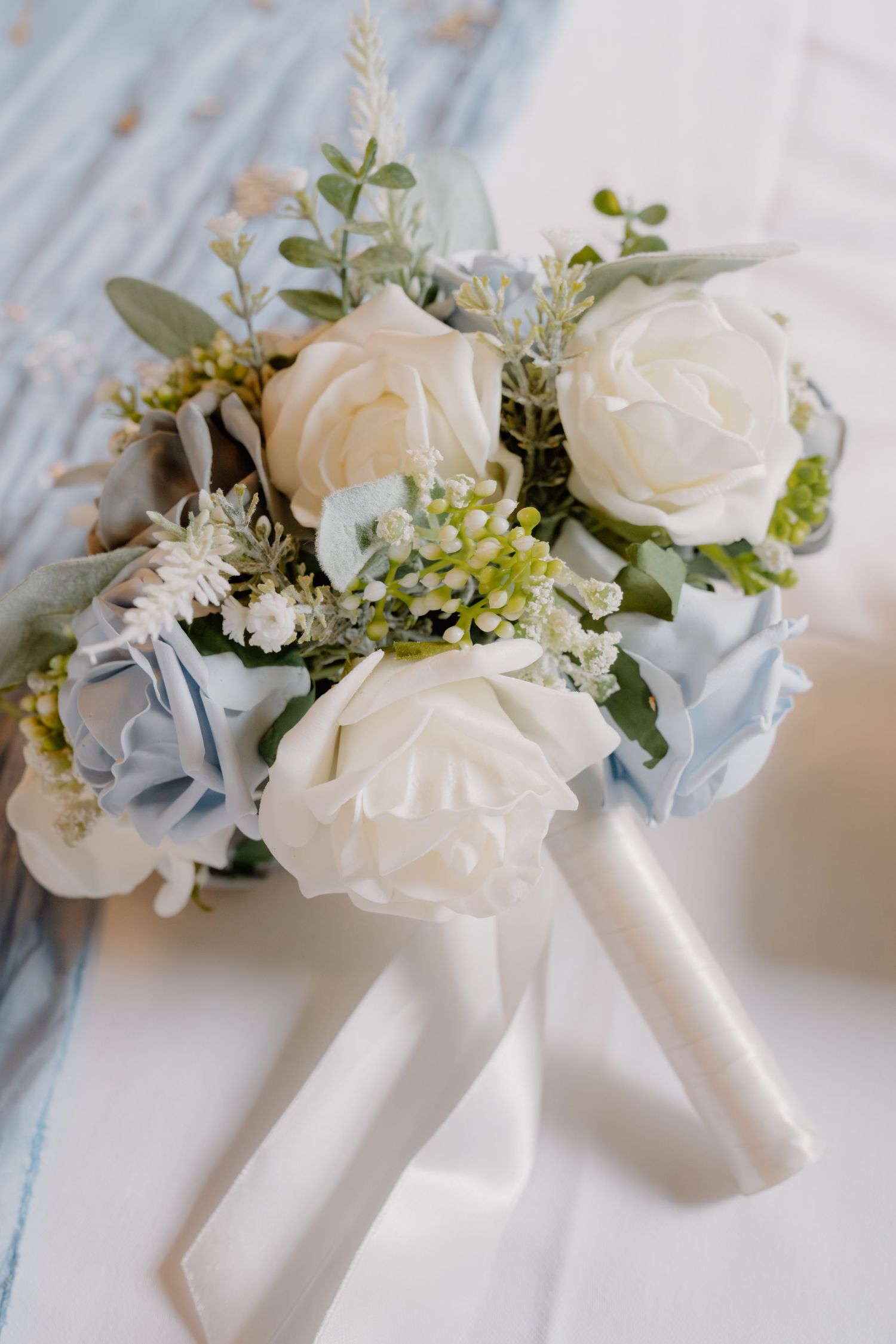 Elegant bridal bouquet with white roses, blue hydrangeas, and greenery, tied with white satin ribbons.