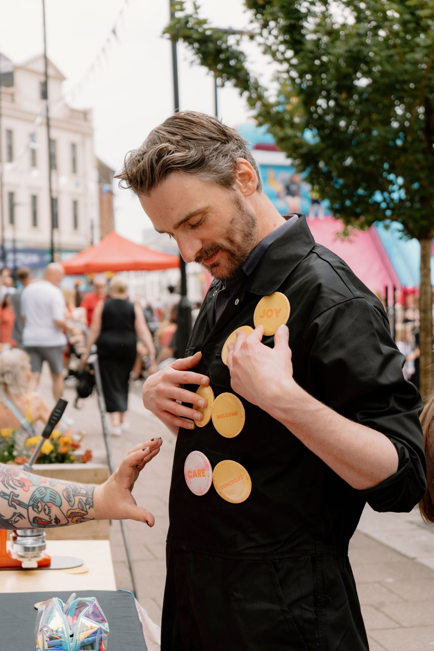 A bearded man in black attire examines golden badges at an outdoor market stall with a tattooed person nearby.