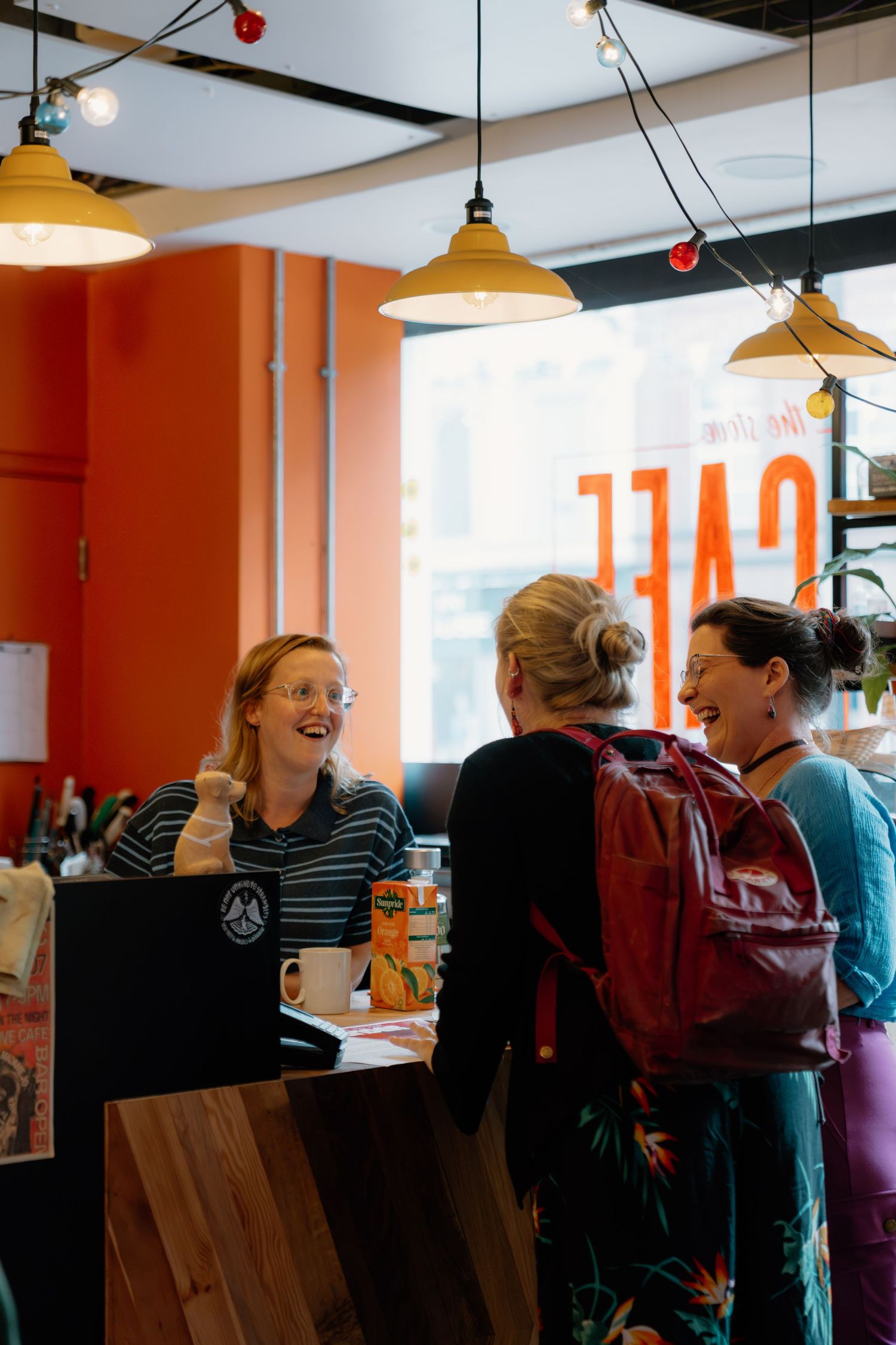 Three women smiling and chatting at a colorful cafe counter with warm pendant lights and orange walls.
