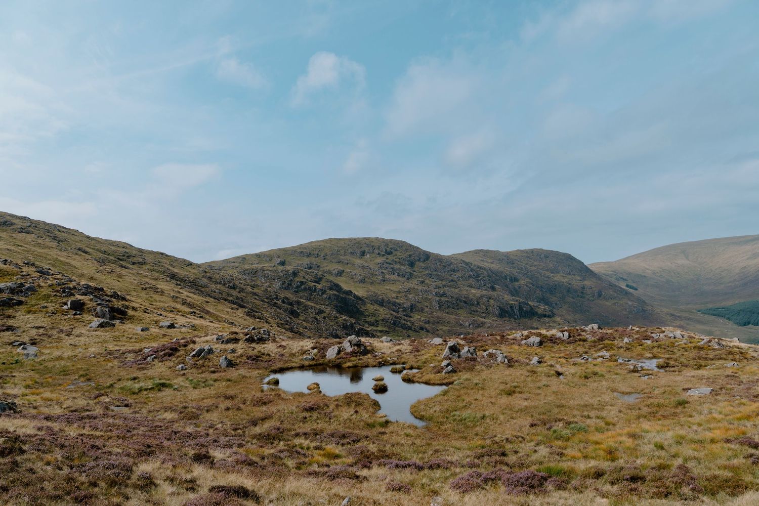 A scenic mountain landscape with small pools of water scattered among rocky terrain under a blue sky with wispy clouds.