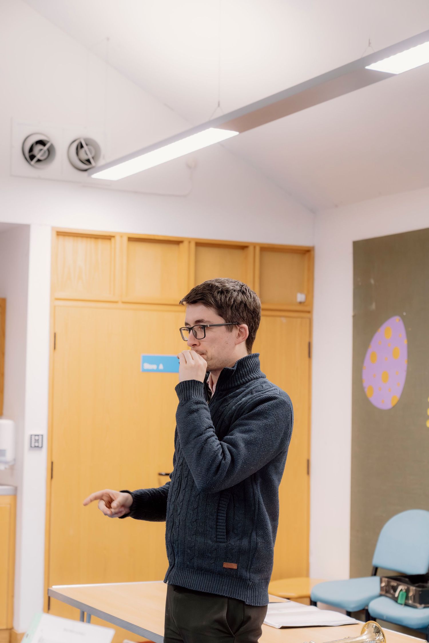 Young person in glasses and gray sweater pointing while standing in a classroom setting.