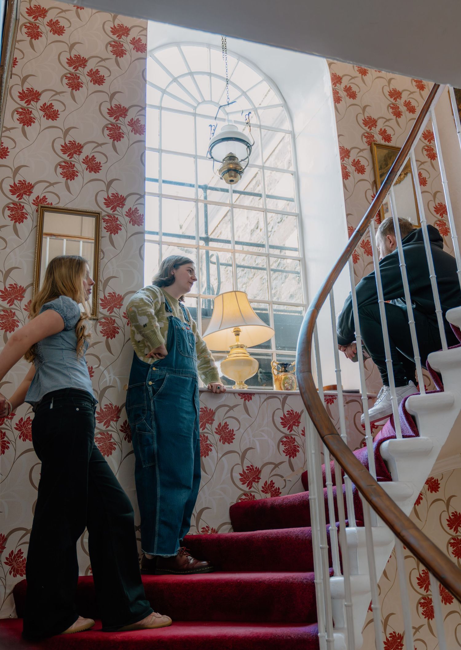 People standing on a red-carpeted staircase in a room with floral wallpaper and a large arched window.