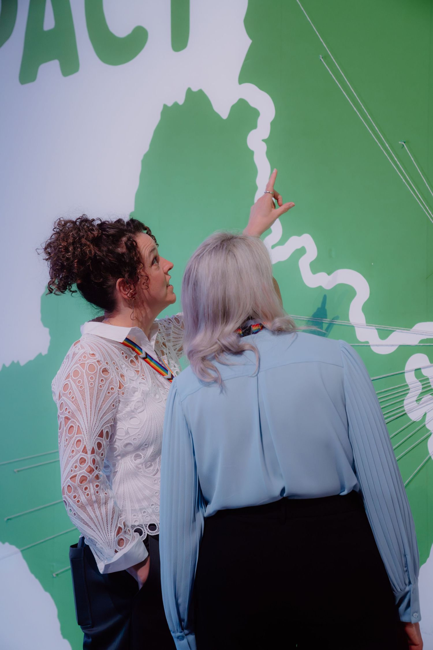 Two women examining a green world map display, one pointing at a location while wearing a rainbow pin.