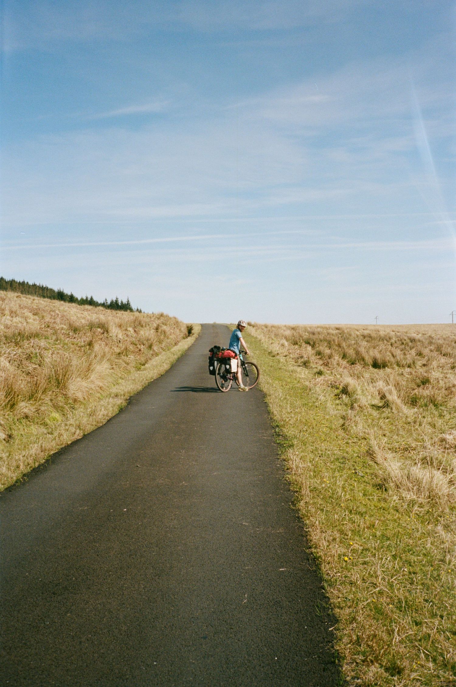 Cyclist with panniers on a narrow rural road through open moorland under a blue sky.