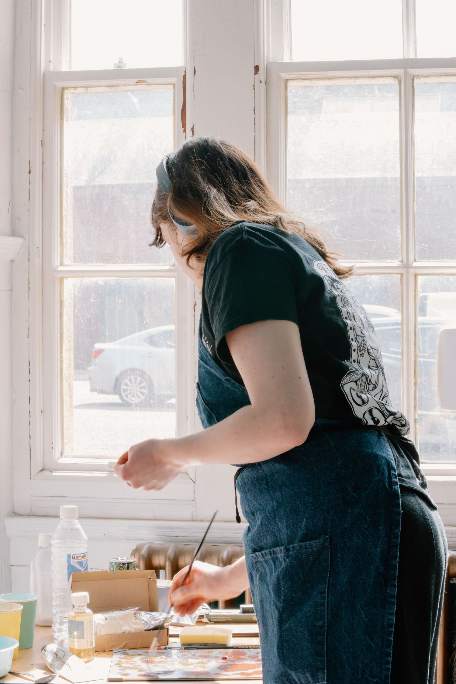 Artist in black t-shirt and denim apron working at a sunlit studio table with art supplies near a bright window.