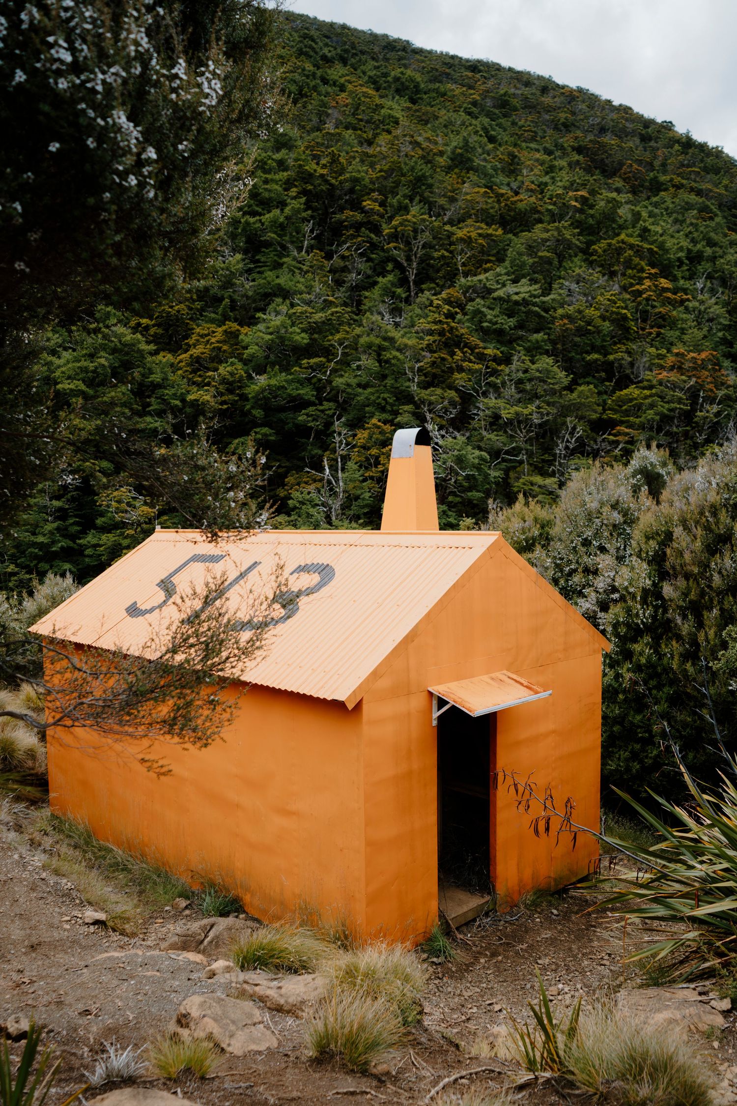 Small orange-painted huts nestled against a hillside with native vegetation in a remote wilderness setting.