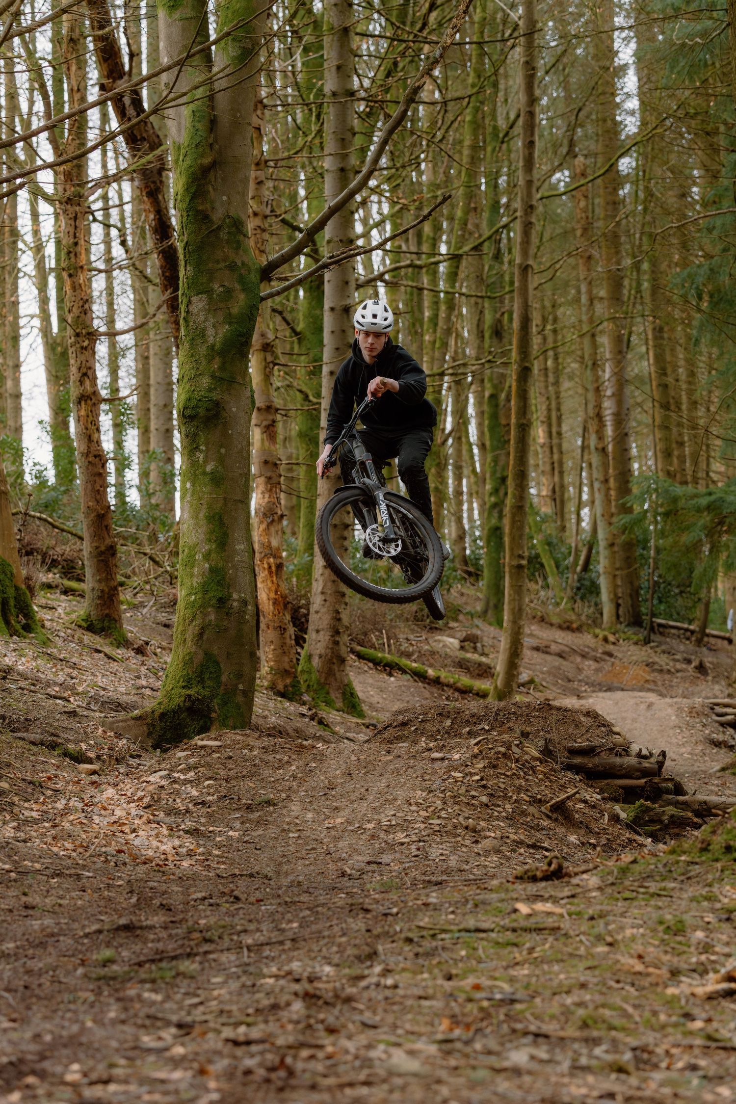 Mountain biker performing a jump trick on a forest trail, wearing a white helmet and dark clothing.