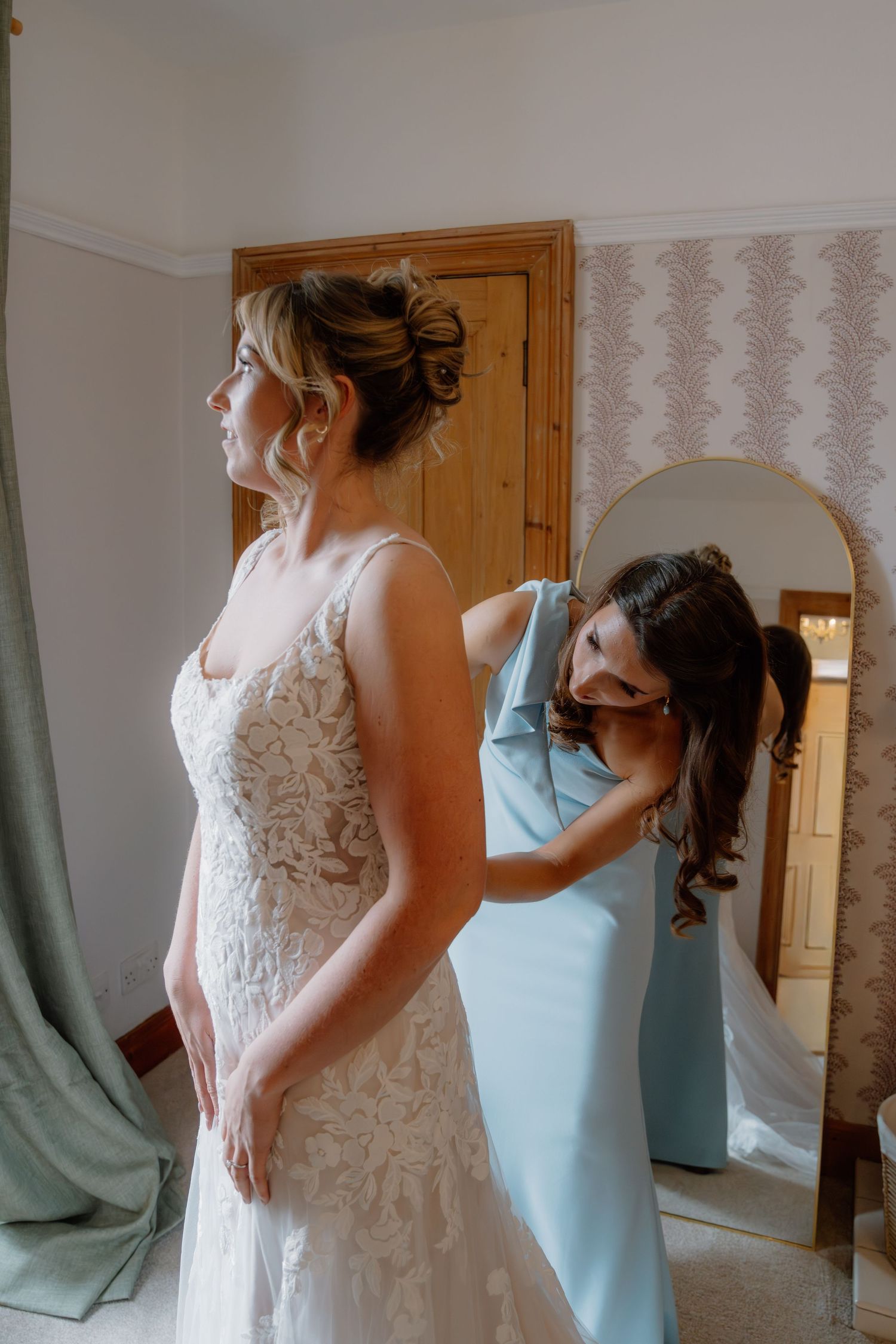 Bridesmaid in light blue dress fastening the back of a bride's lace wedding gown before the ceremony.