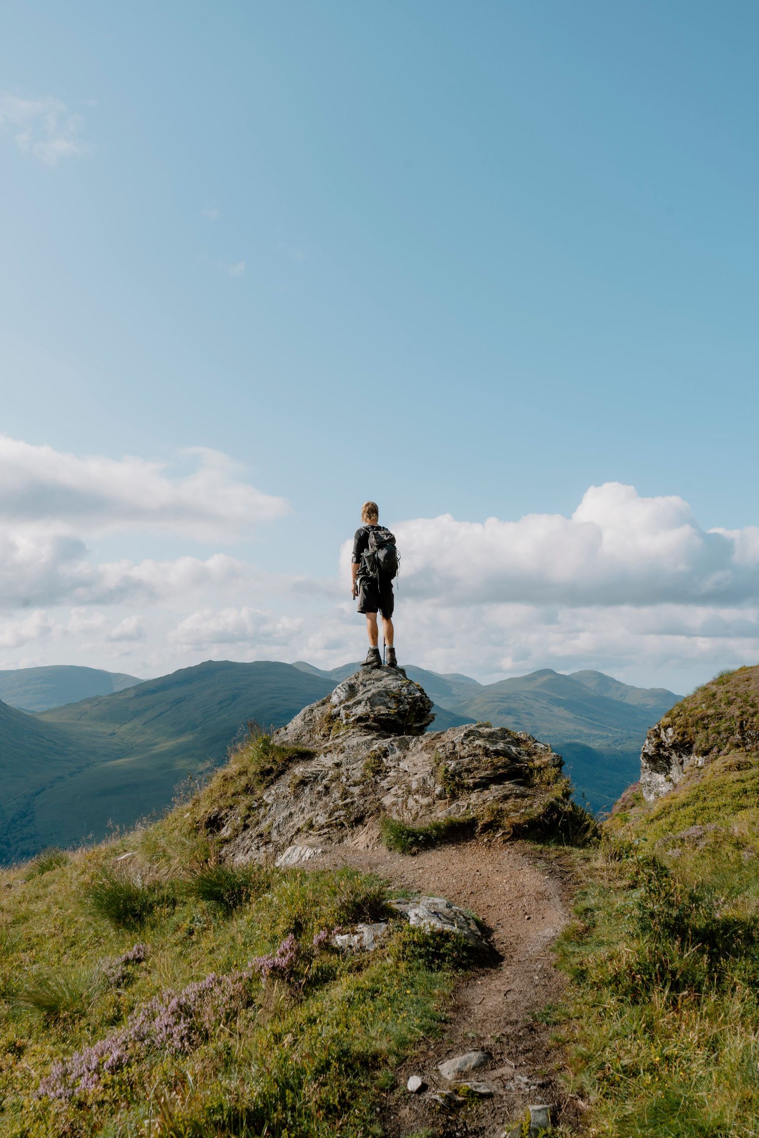 A hiker stands atop a rocky outcrop overlooking rolling mountain peaks under a bright blue sky with scattered clouds.