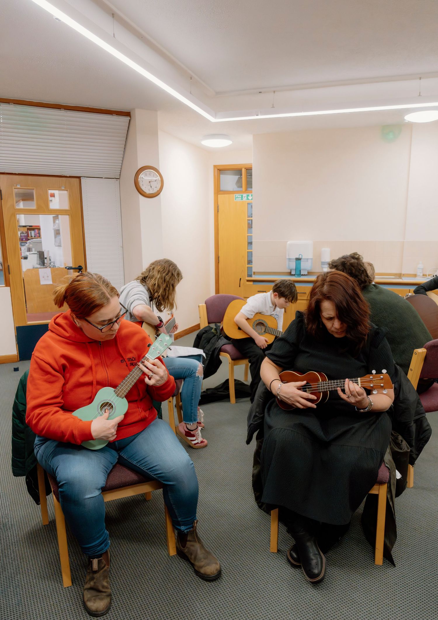 Group of people sitting in a circle playing ukuleles and guitars in a music class session indoors.