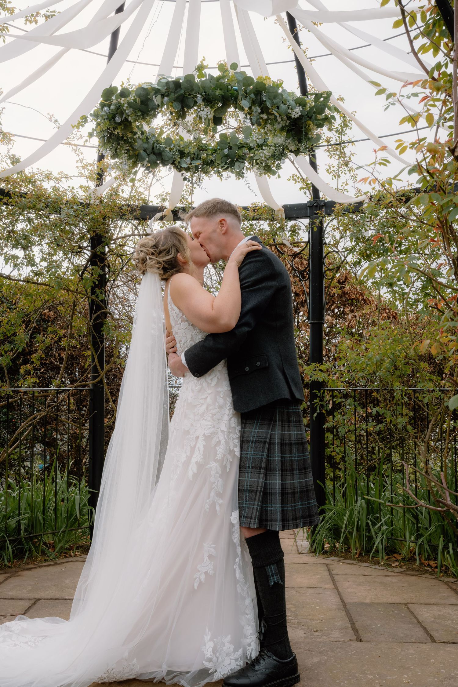 Bride and groom share first kiss under floral arch, groom wearing traditional Scottish tartan kilt, in a garden ceremony.