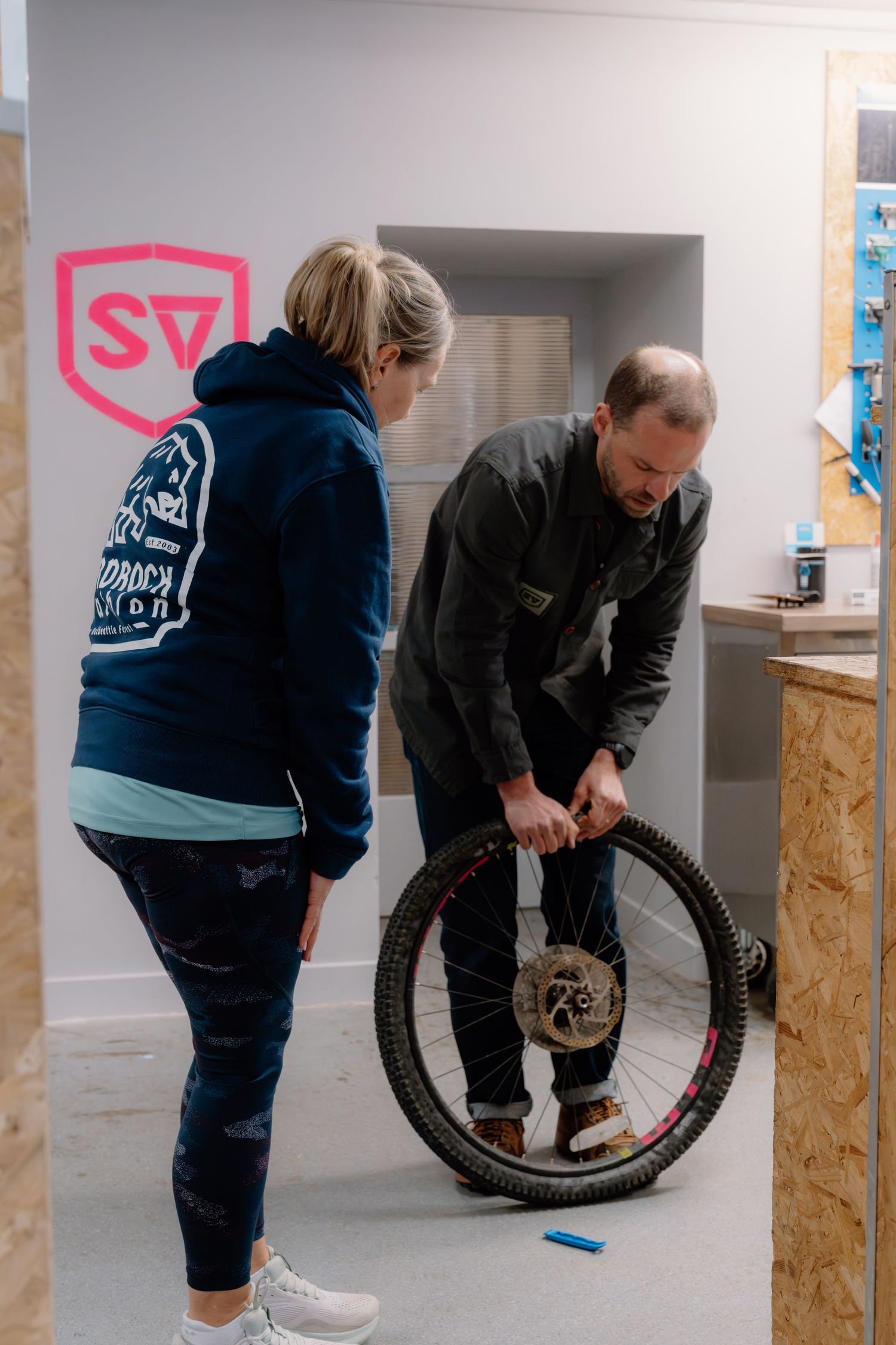 A man repairs a bicycle wheel while a woman in a blue hoodie watches inside a bike shop.