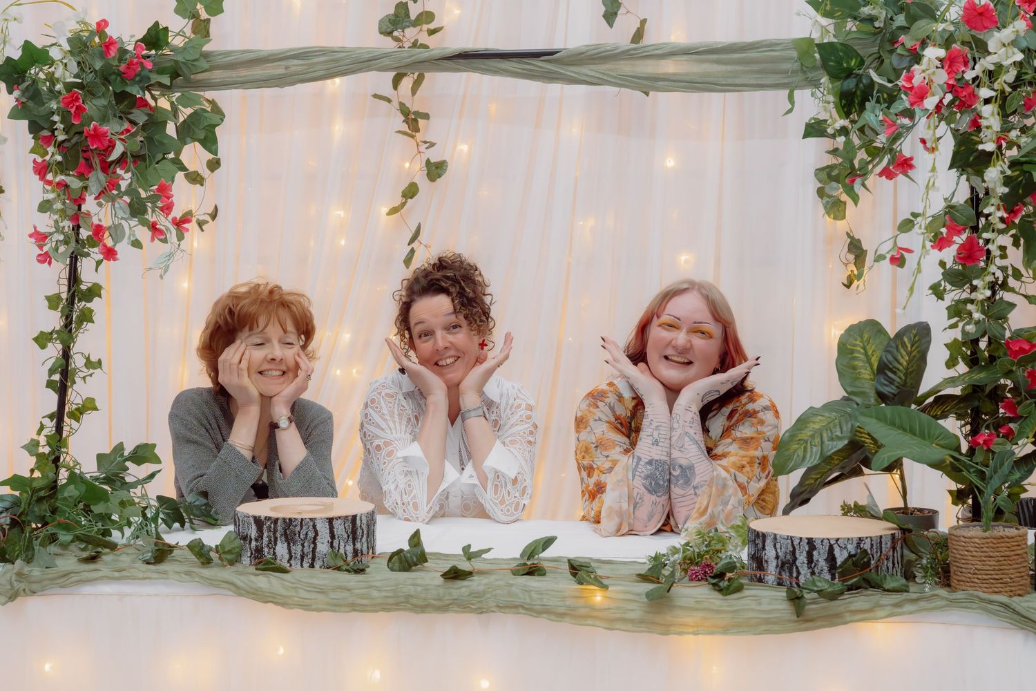 Three smiling women pose together at a decorated table with fairy lights, greenery, and pink flowers backdrop.