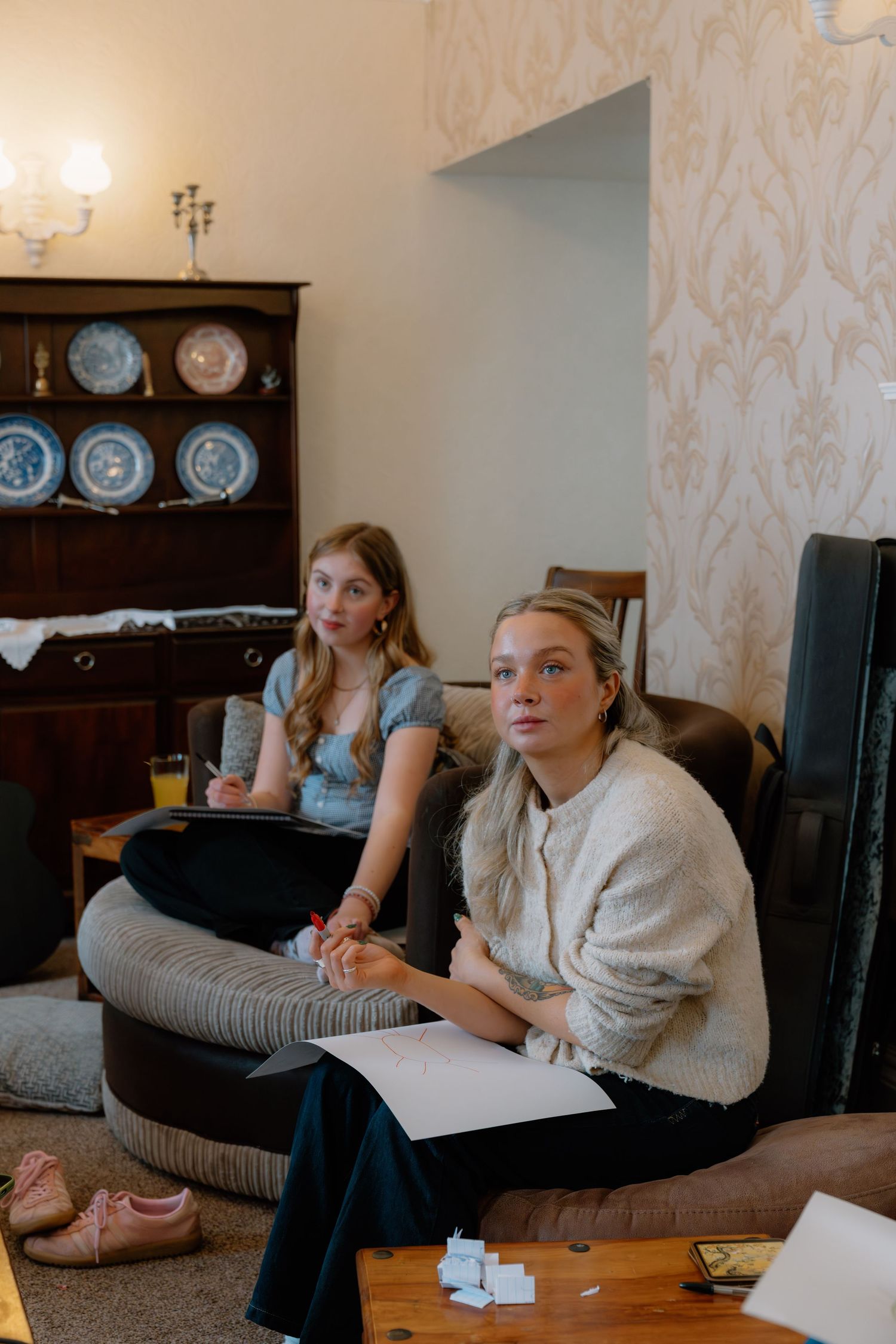 Two young women sitting on sofas in a cozy living room, attentively watching something while holding papers.
