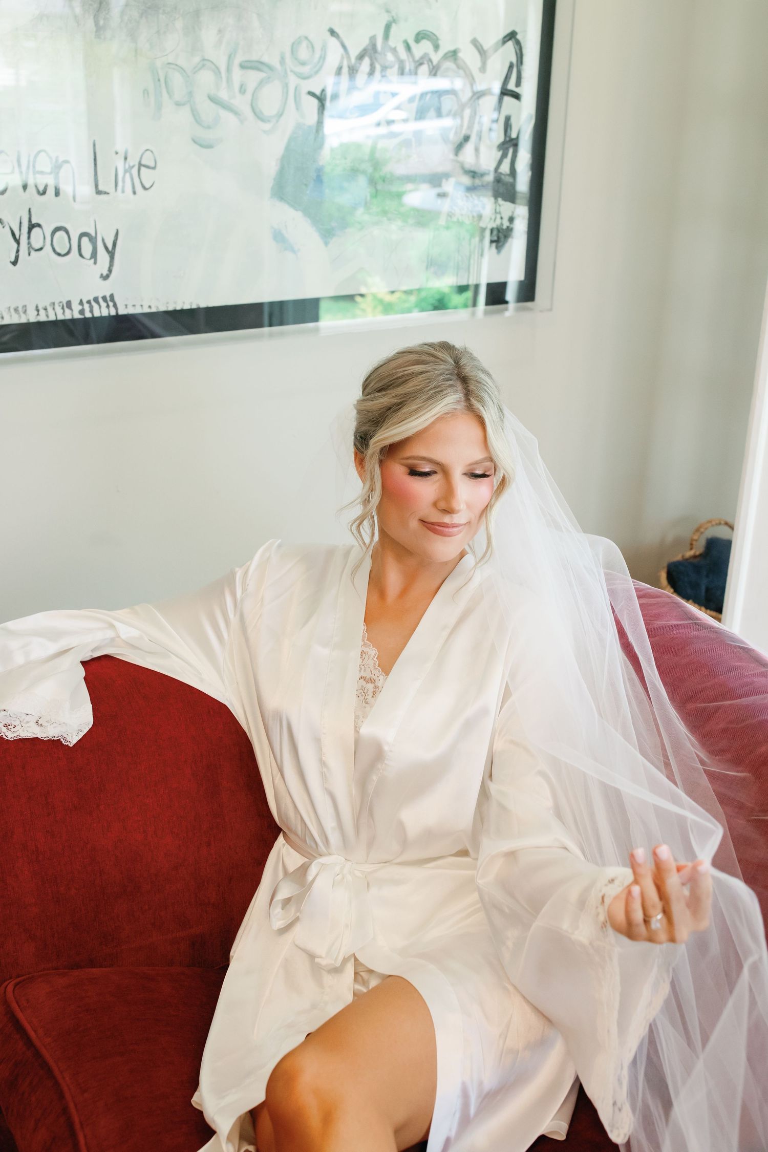 Bride in white satin robe and veil sits on red velvet sofa, admiring her engagement ring before wedding.