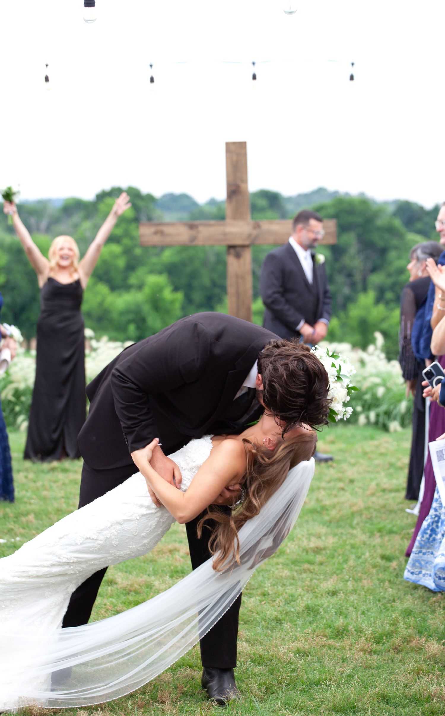 Groom dips bride for a kiss at outdoor wedding ceremony with wooden cross and cheering wedding party.