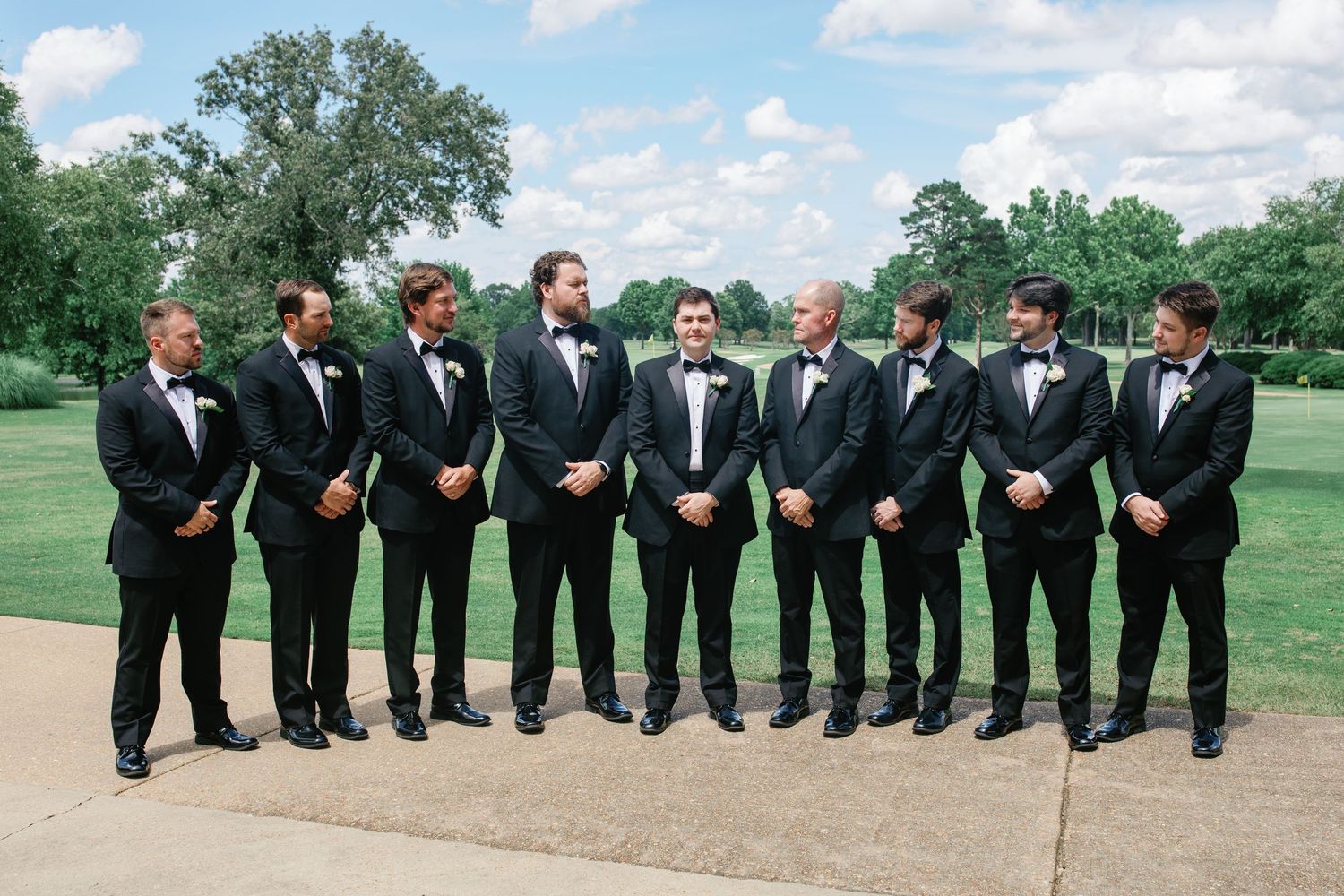 Groom and groomsmen in black tuxedos with white boutonnieres posing outdoors on a sunny wedding day.