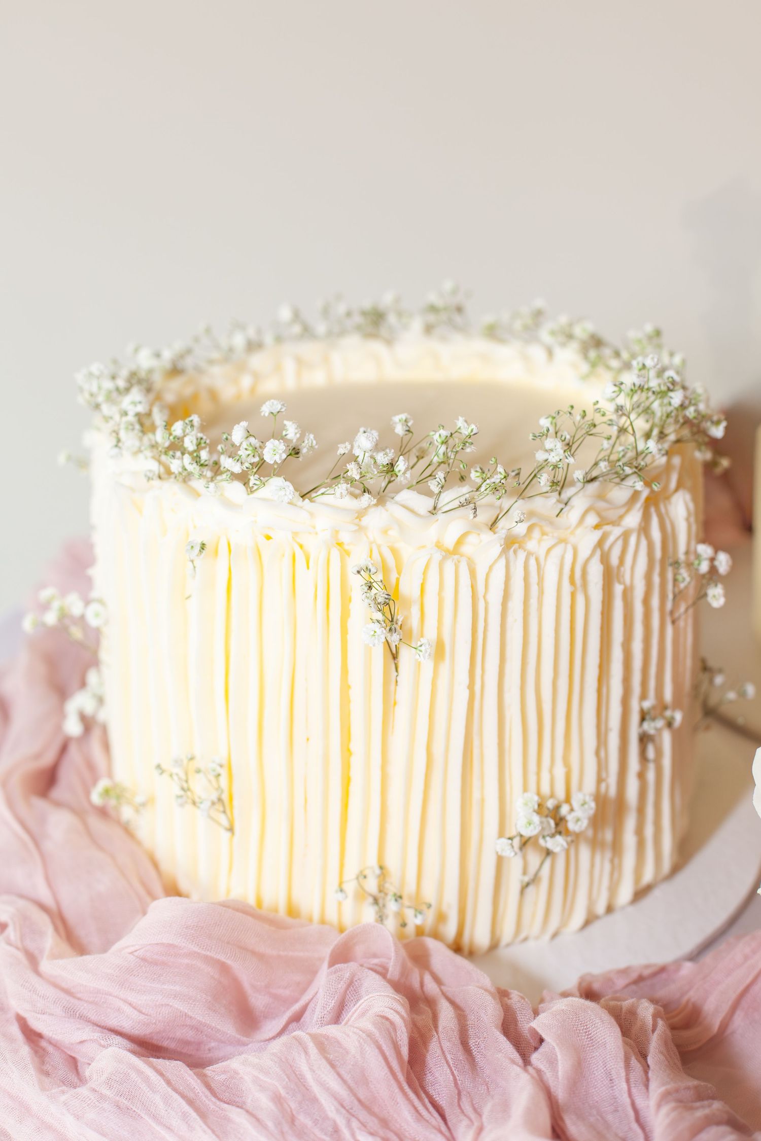 Elegant white cake with drip frosting and delicate baby's breath flowers on top, displayed on pink fabric.