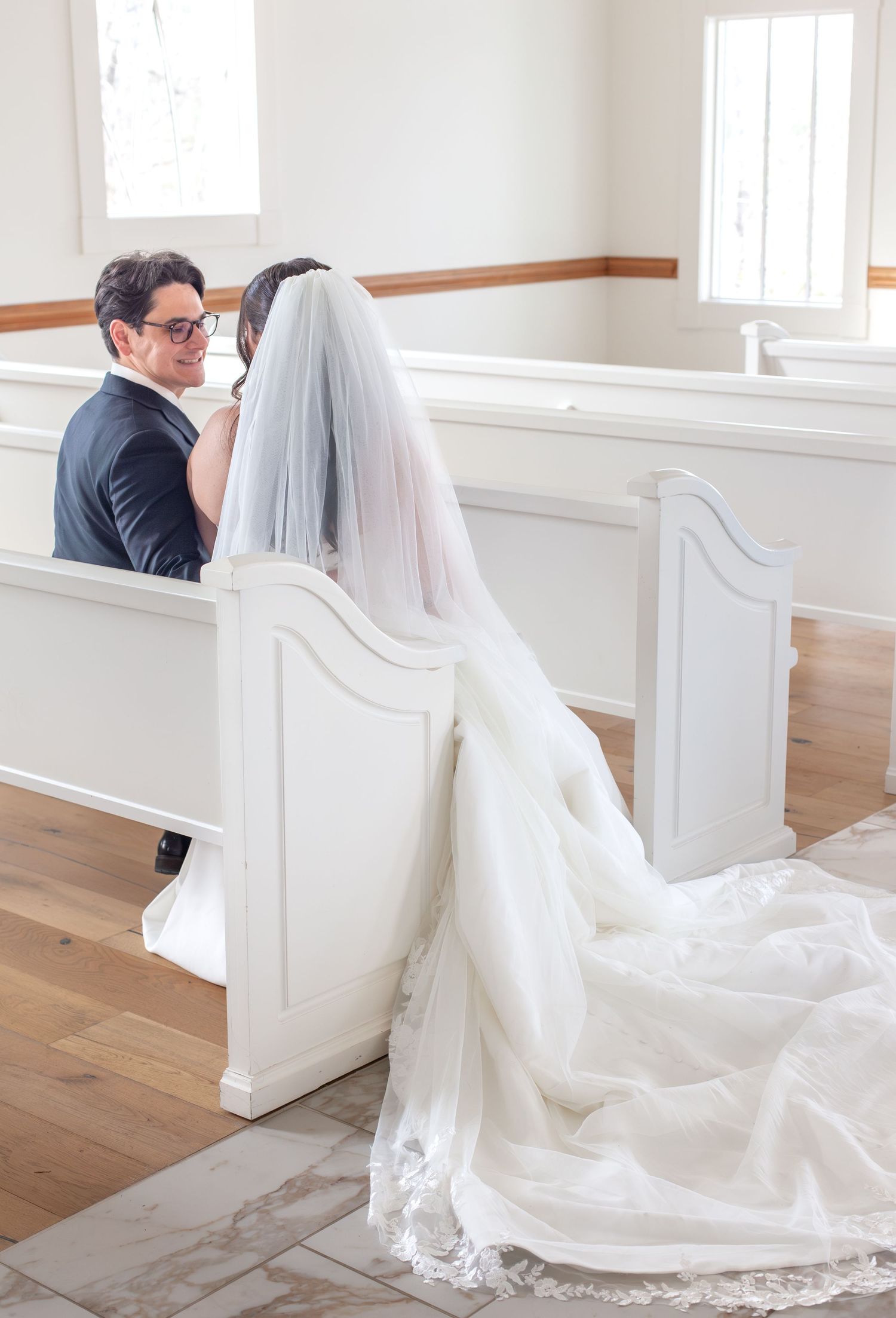 A bride in a white gown and veil sits with a groom in a dark suit on white church pews during their wedding ceremony.