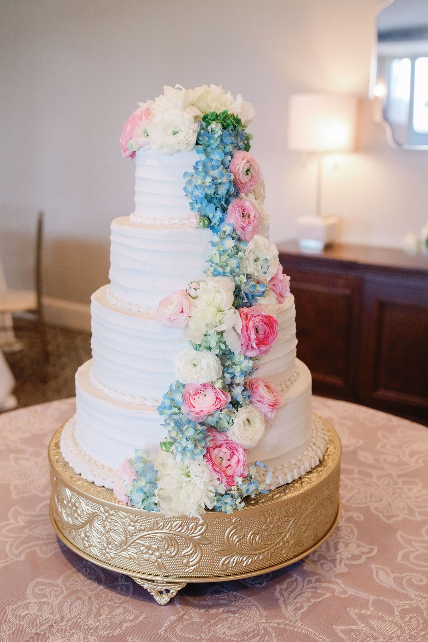 Four-tier white wedding cake with pink roses and blue hydrangeas on a gold stand.