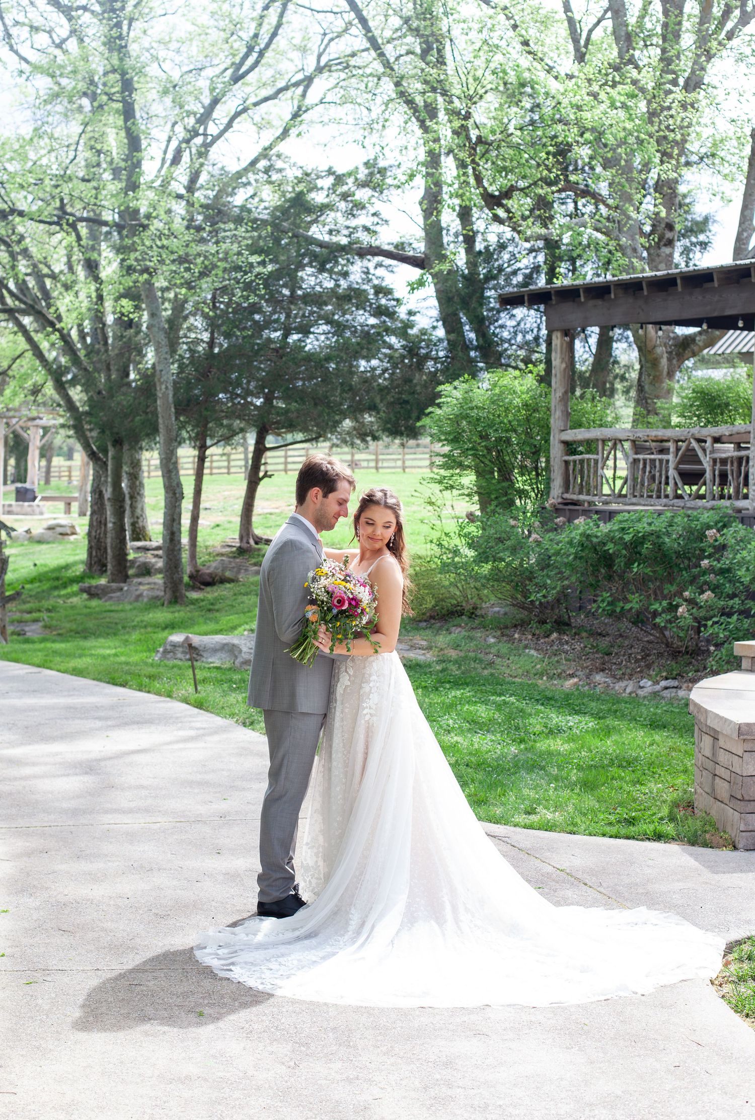 Bride in white gown holding colorful bouquet stands with groom in gray suit in lush garden setting.