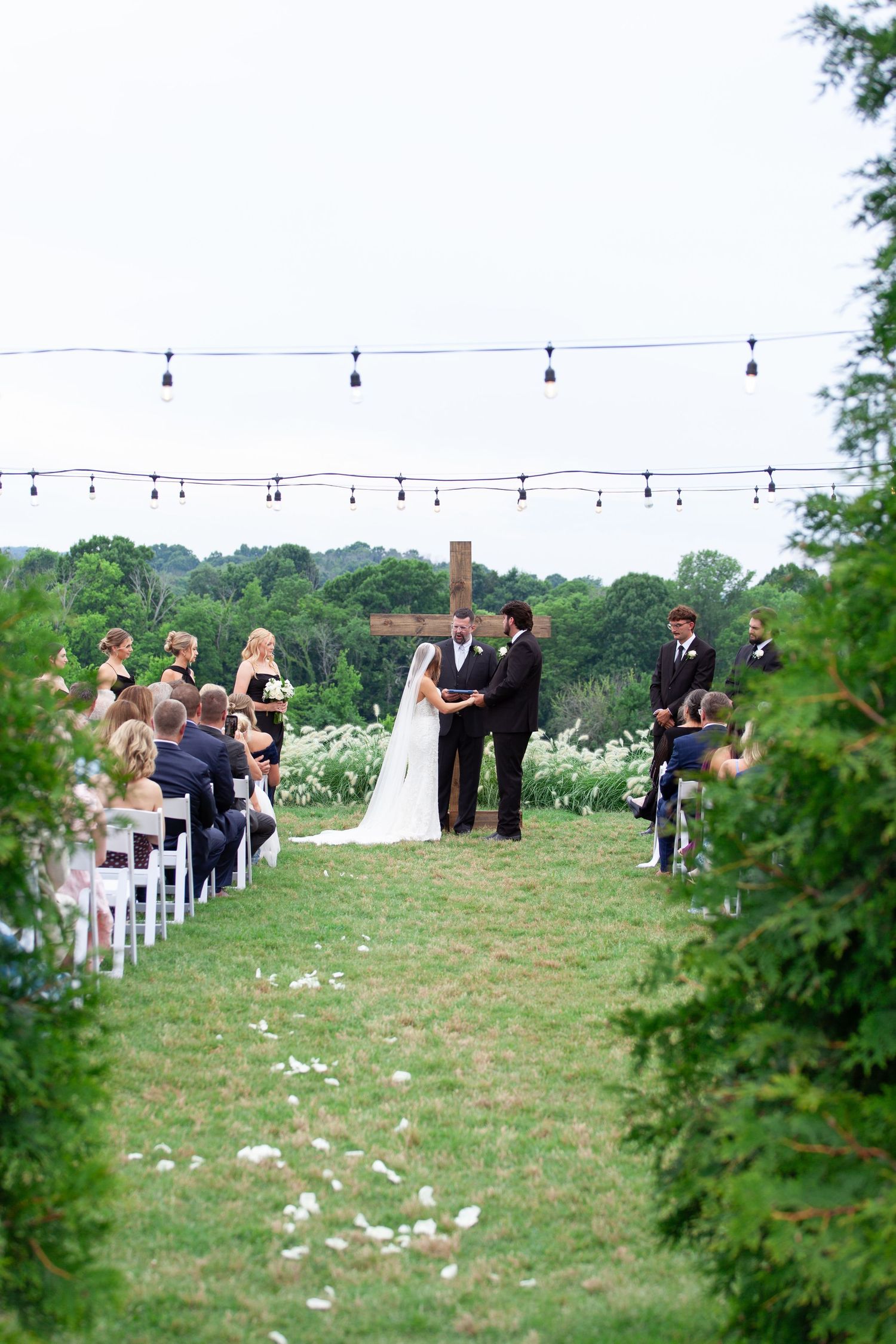 Outdoor wedding ceremony on green lawn with couple at altar, wooden cross, string lights, and guests seated in white chairs.