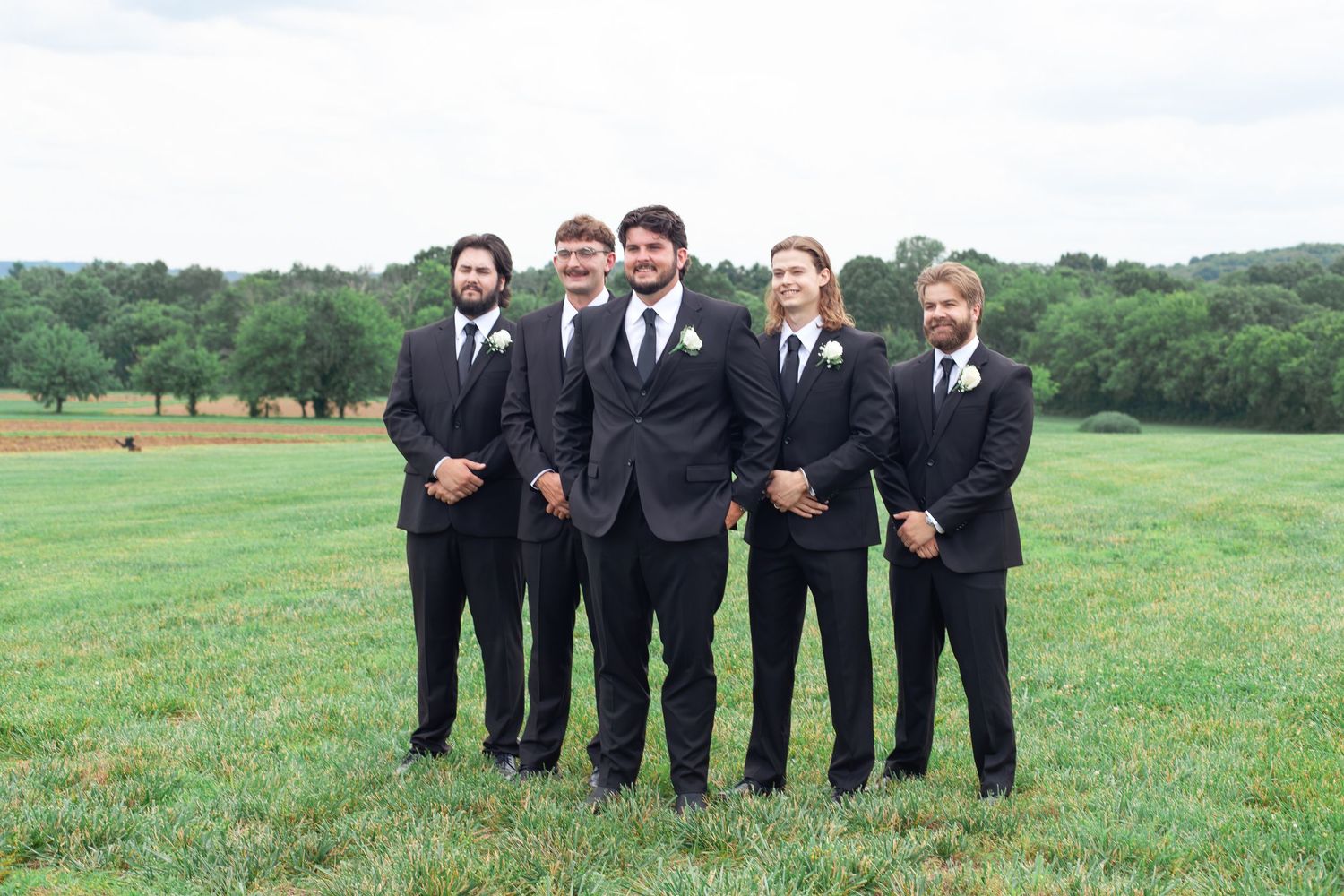 Five groomsmen in black suits pose outdoors on a green field with trees in the background at a wedding.