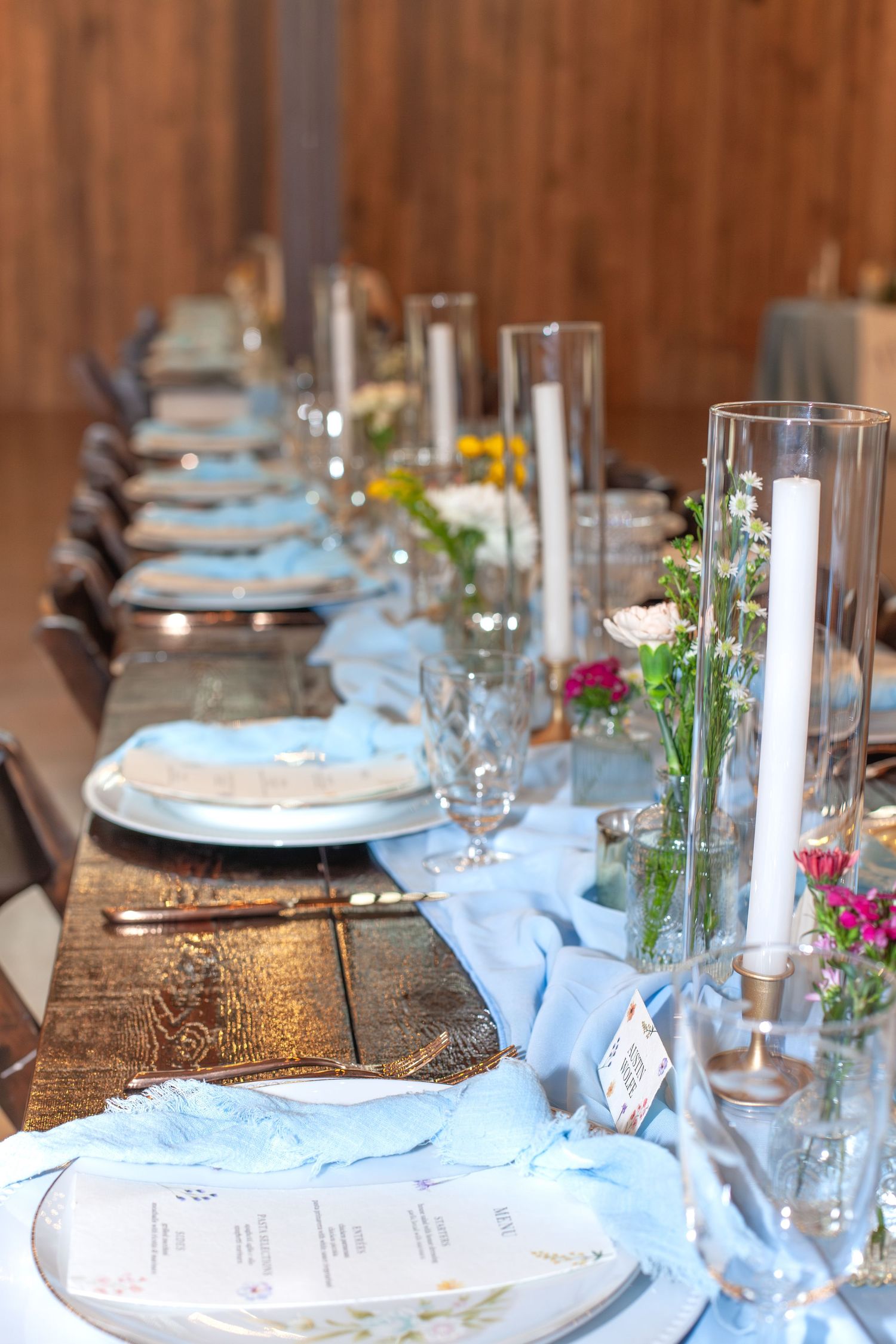 Elegant wedding reception table set with blue runner, white candles, floral arrangements, and place settings on rustic wood.