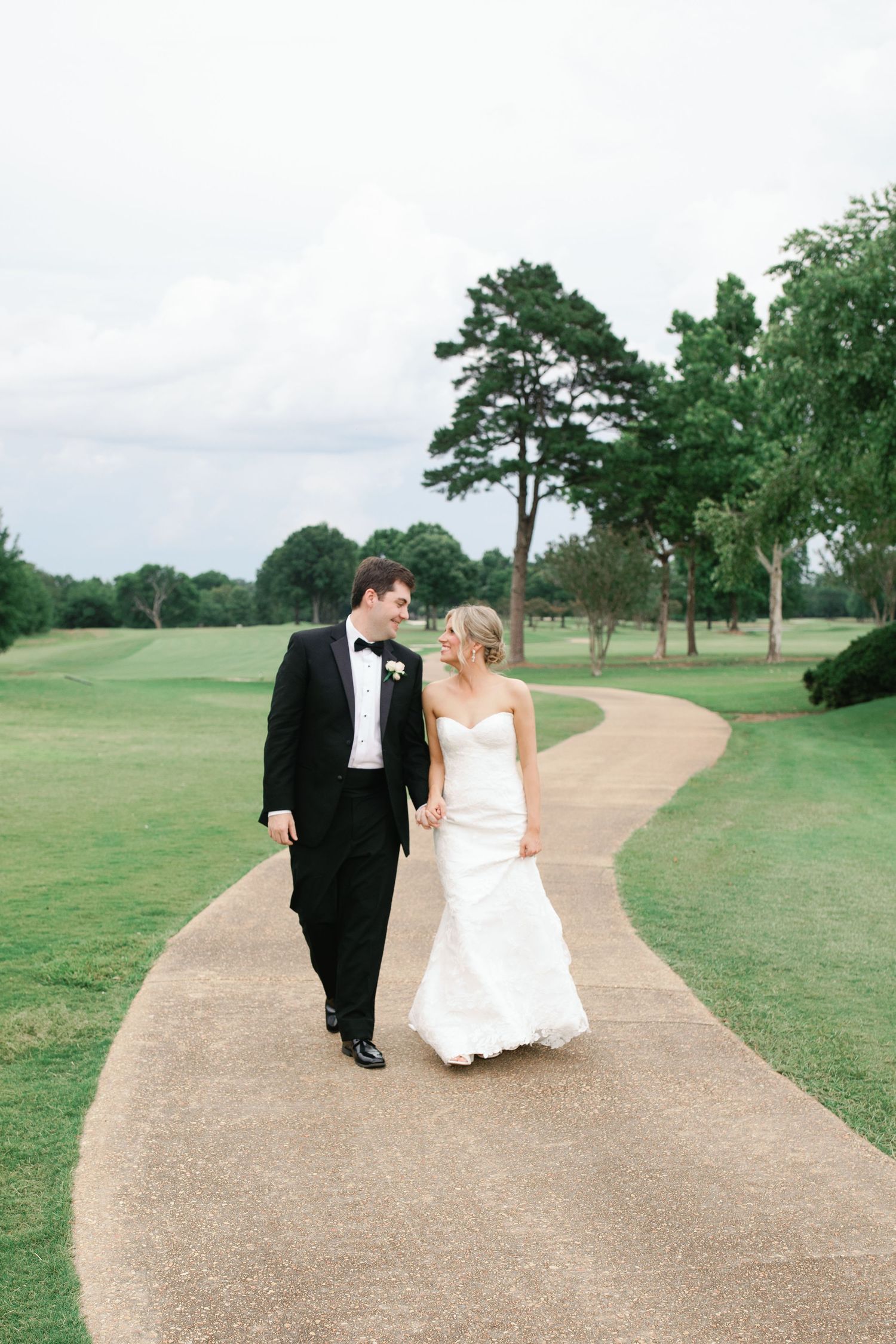 Bride and groom walk hand-in-hand along a winding path at a lush green golf course on their wedding day.
