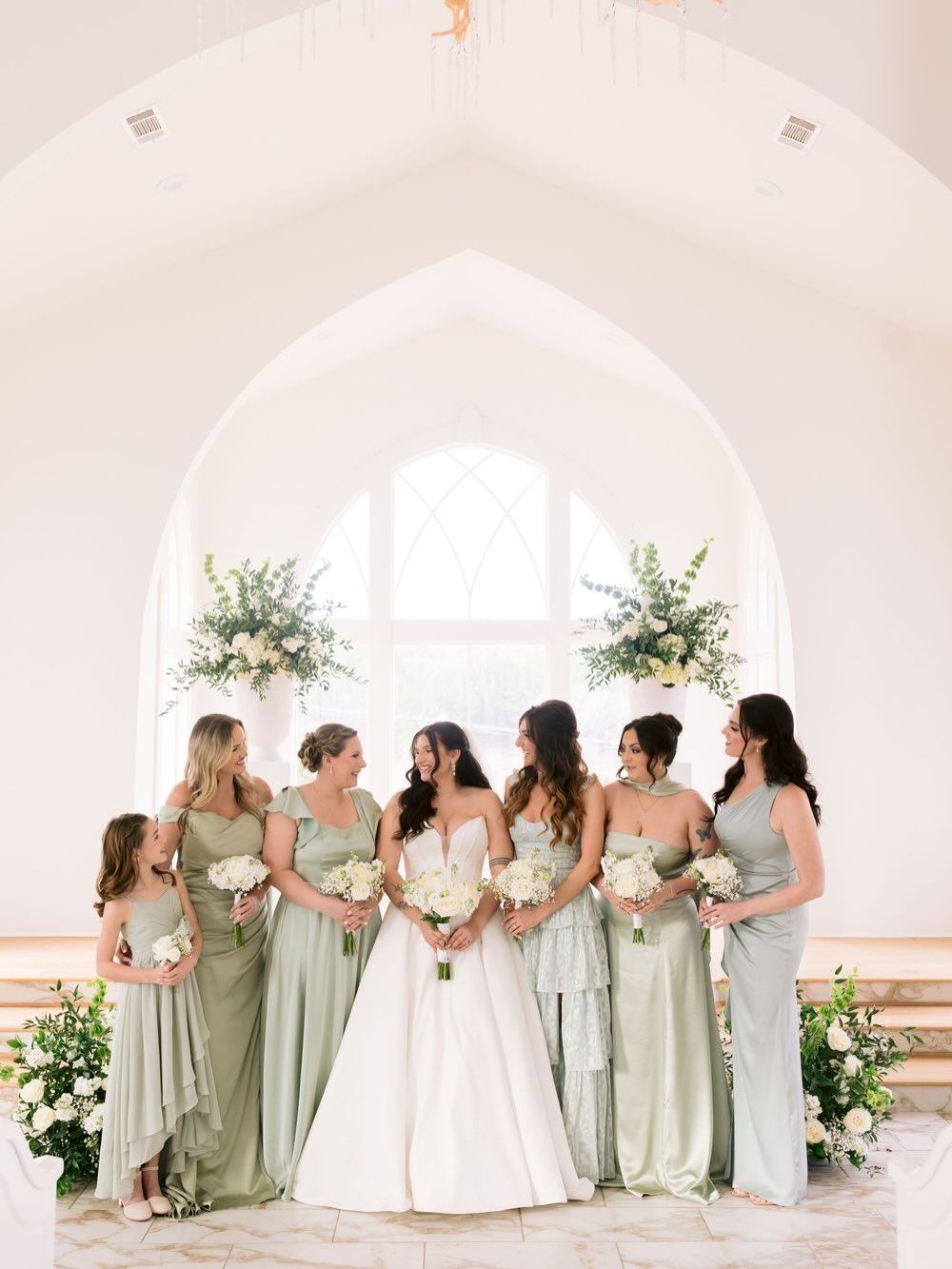 A bridal party in sage green bridesmaid dresses and white wedding gown pose together in an elegant white venue.