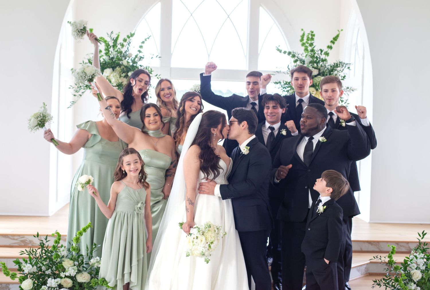 Wedding party celebrates as bride and groom kiss at church altar, bridesmaids in sage green gowns, groomsmen in black suits.