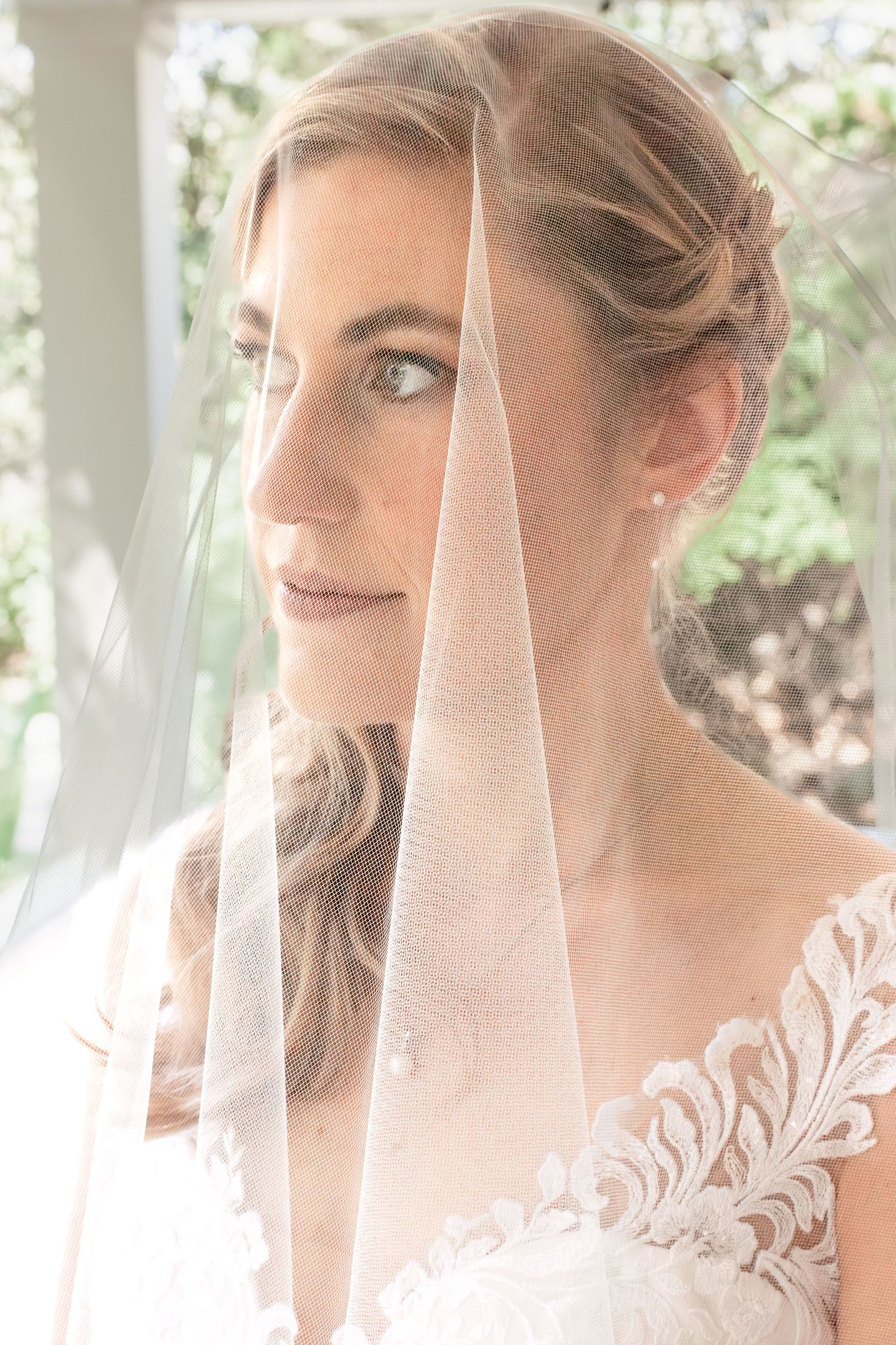 A bride in a delicate lace wedding dress with a sheer veil poses in soft natural lighting.