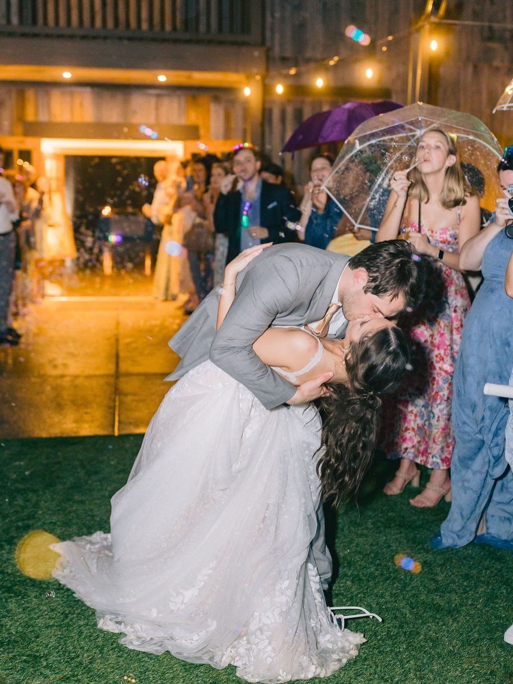 Bride and groom share a romantic dip kiss outdoors at night, surrounded by cheering wedding guests with umbrellas.
