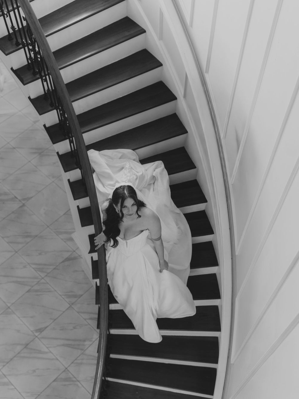 Black and white aerial view of a bride in a white gown posing gracefully on a curved staircase.