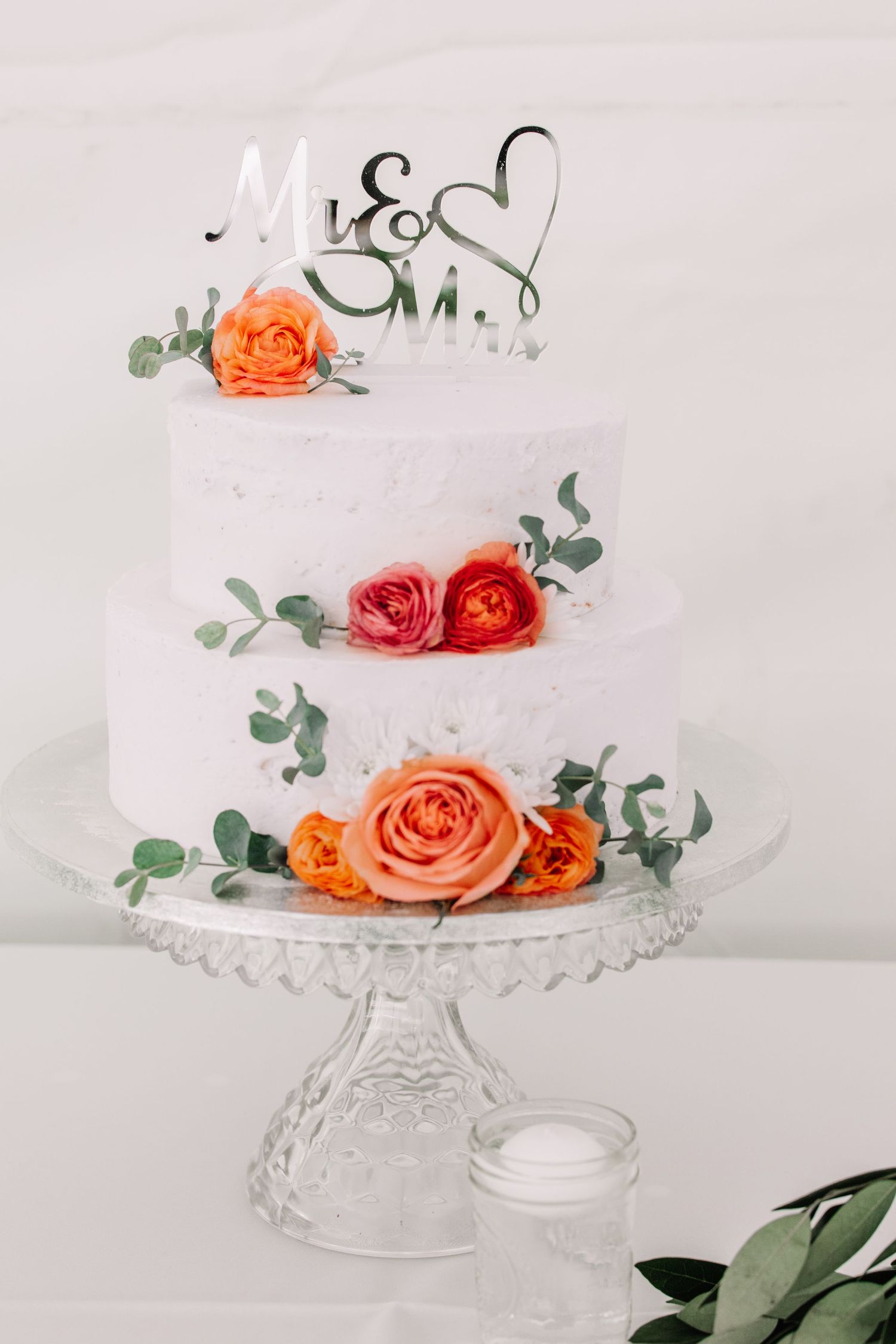 An elegant white wedding cake decorated with coral roses and eucalyptus on a crystal stand.