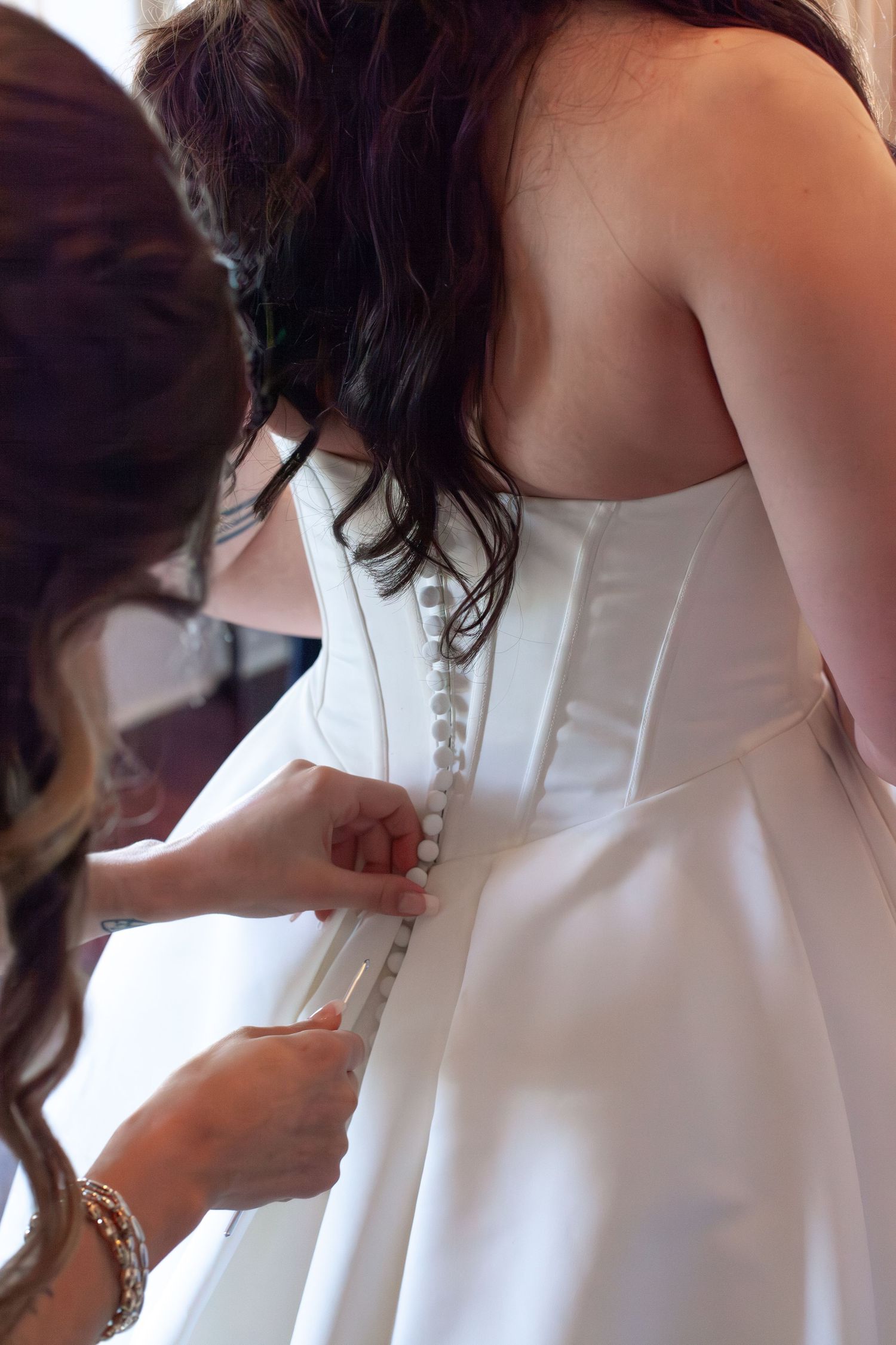 Bridesmaid fastening pearl buttons on the back of a white satin wedding dress.